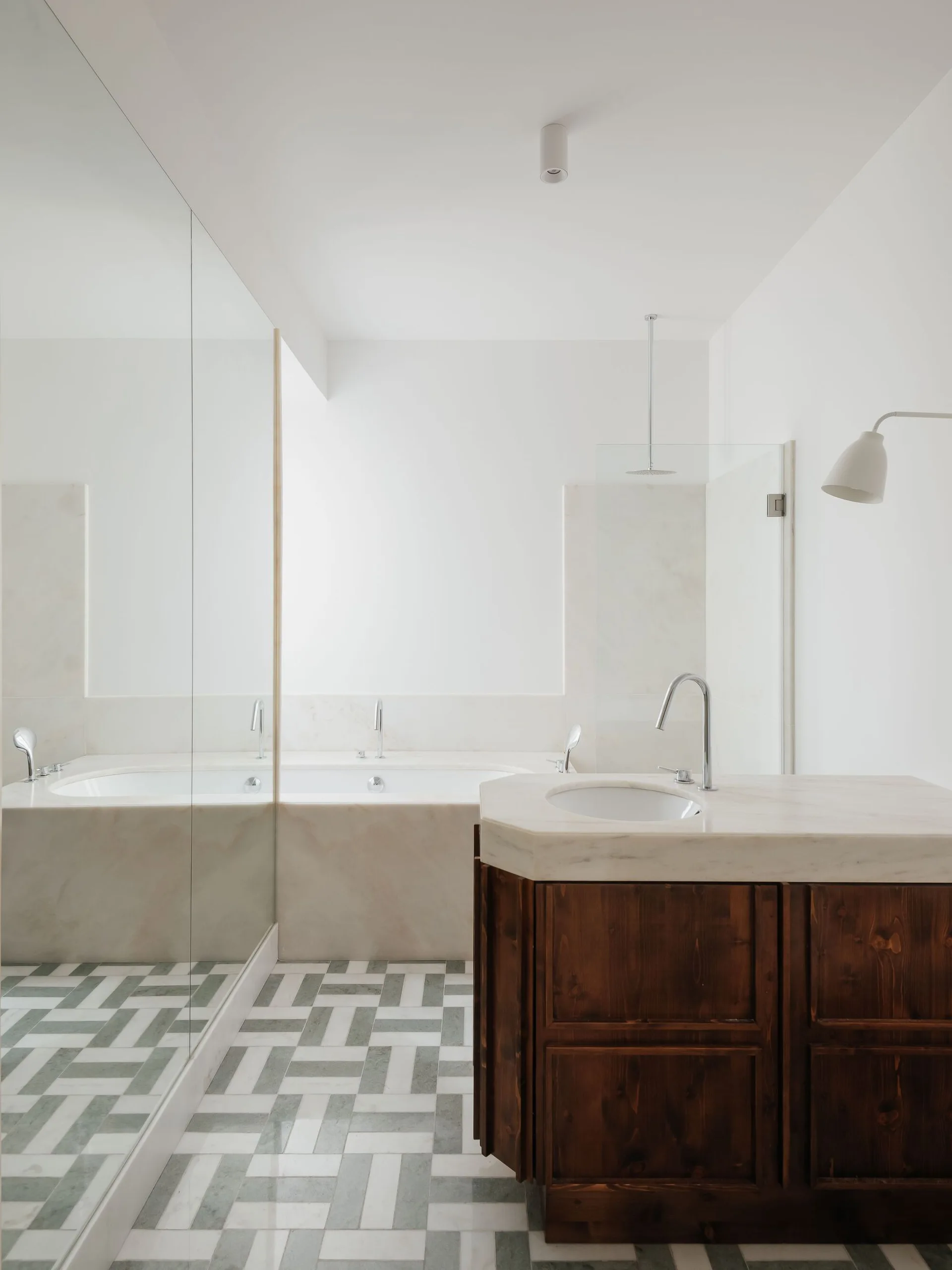 A minimalist bathroom features a dark wood vanity with a marble countertop, a built-in bathtub, and a grey and white geometric tiled floor.