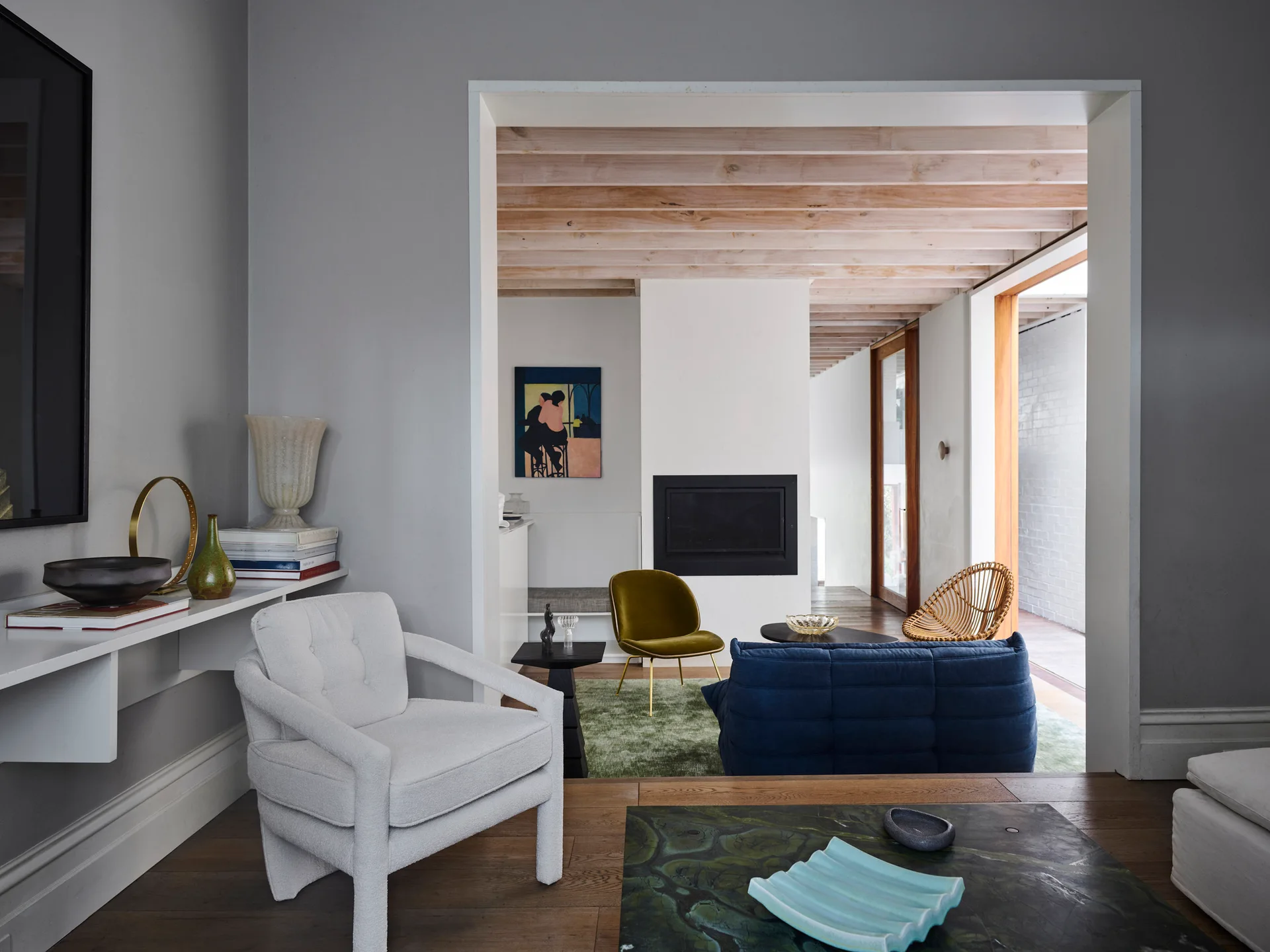 A modern living room viewed through an archway, featuring a blue sofa, white armchair, fireplace, and exposed wood ceiling beams.