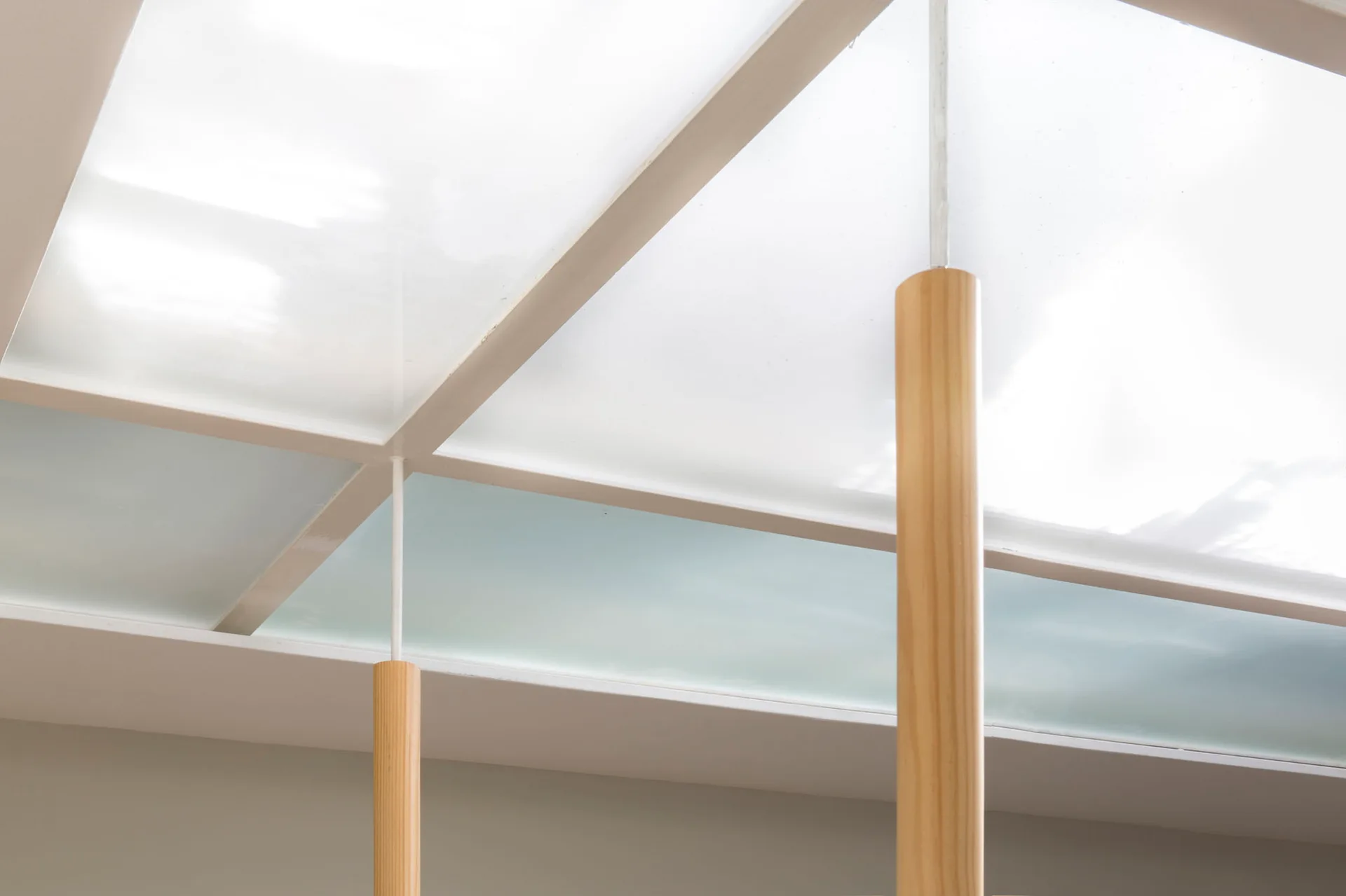 A low-angle view of a geometric white-framed skylight with frosted glass supported by two vertical, cylindrical light wood columns.