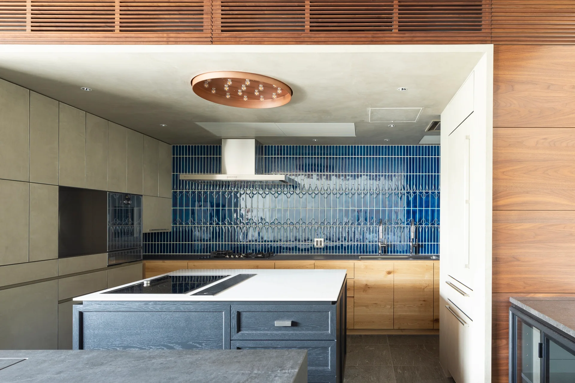 A modern kitchen featuring a glossy blue vertical tile backsplash, wood cabinets, and a grey kitchen island with a white countertop.