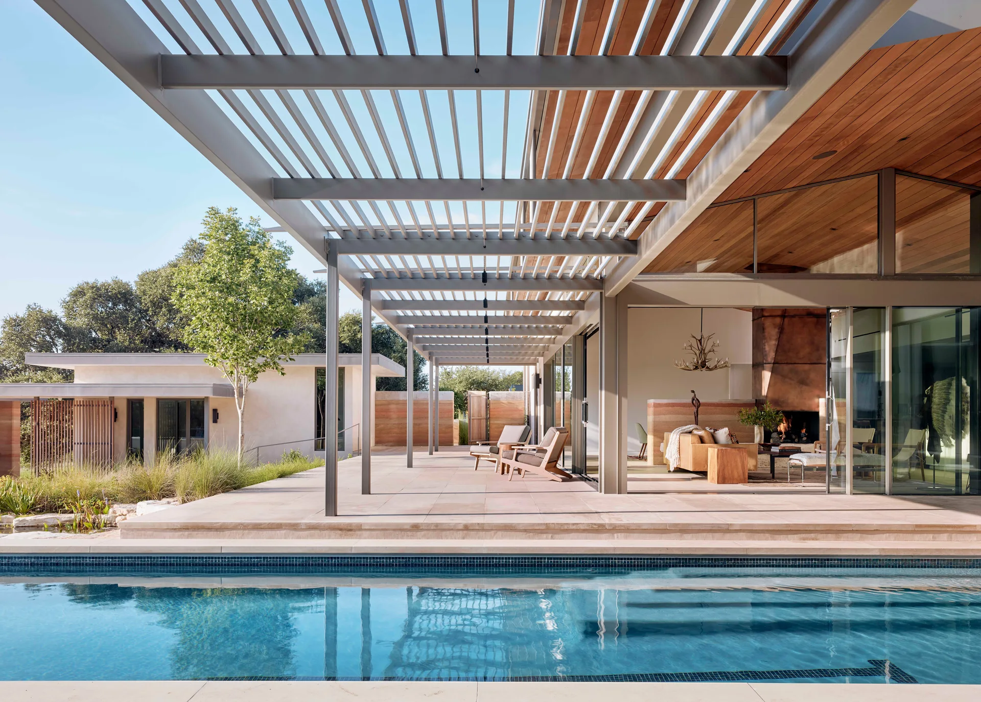 A rectangular swimming pool sits next to a stone tile patio covered by a metal pergola, with lounge chairs and sliding glass doors leading indoors.