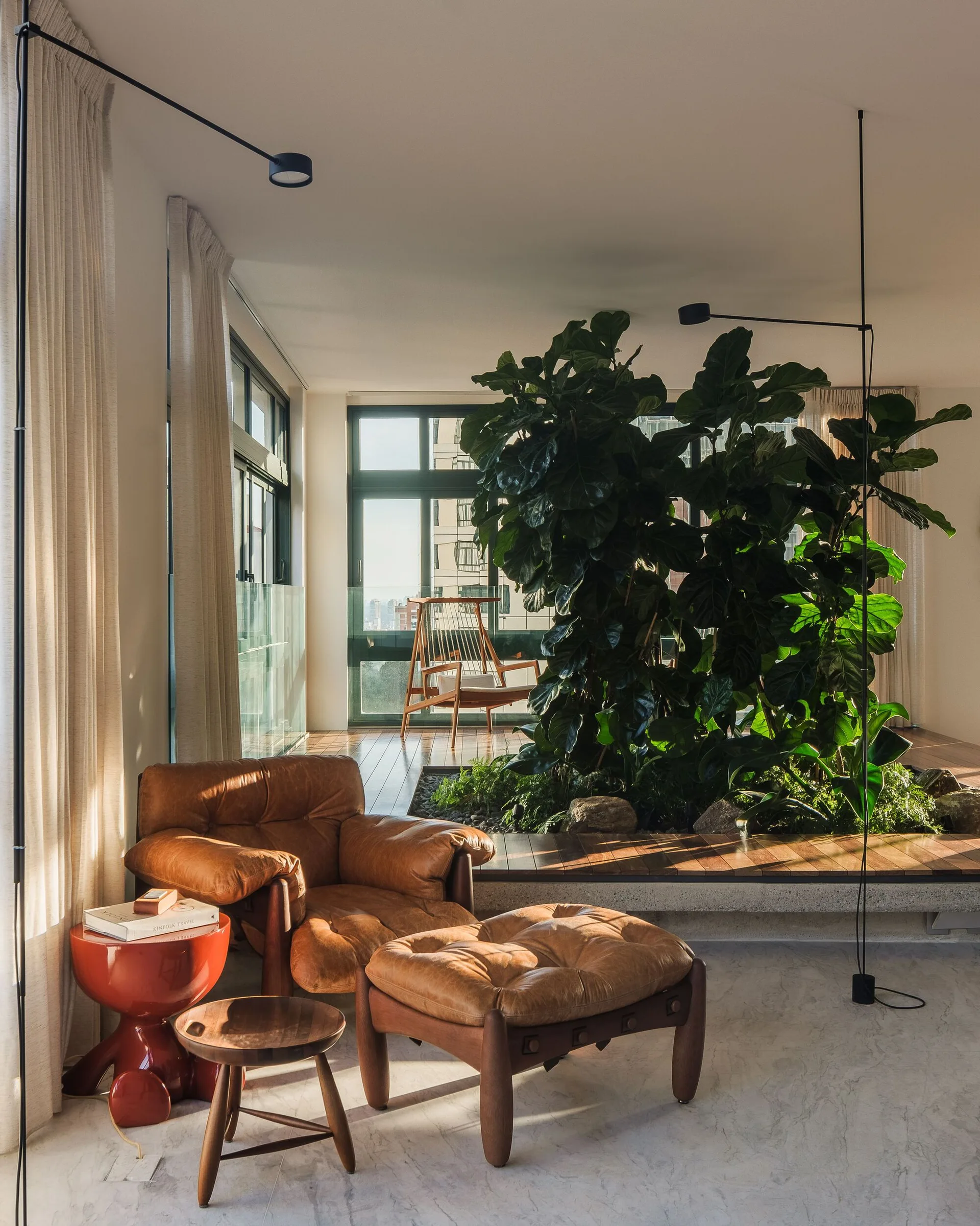 A brown leather lounge chair and ottoman sit beside a sunken indoor garden with a large green tree in a sunlit room.
