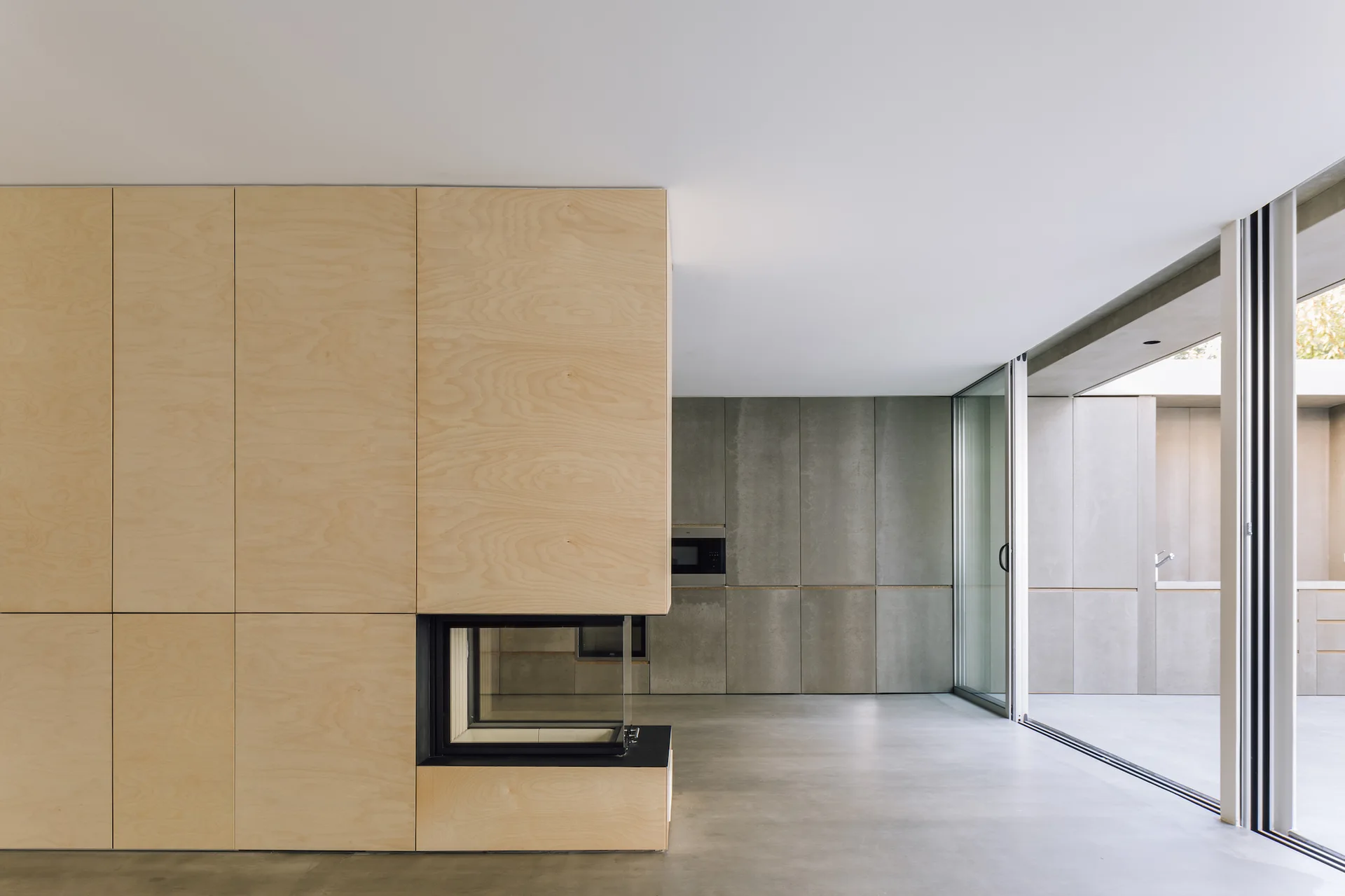 A modern minimalist space featuring a light plywood storage wall with a built-in corner fireplace, gray concrete cabinets, and a concrete floor.