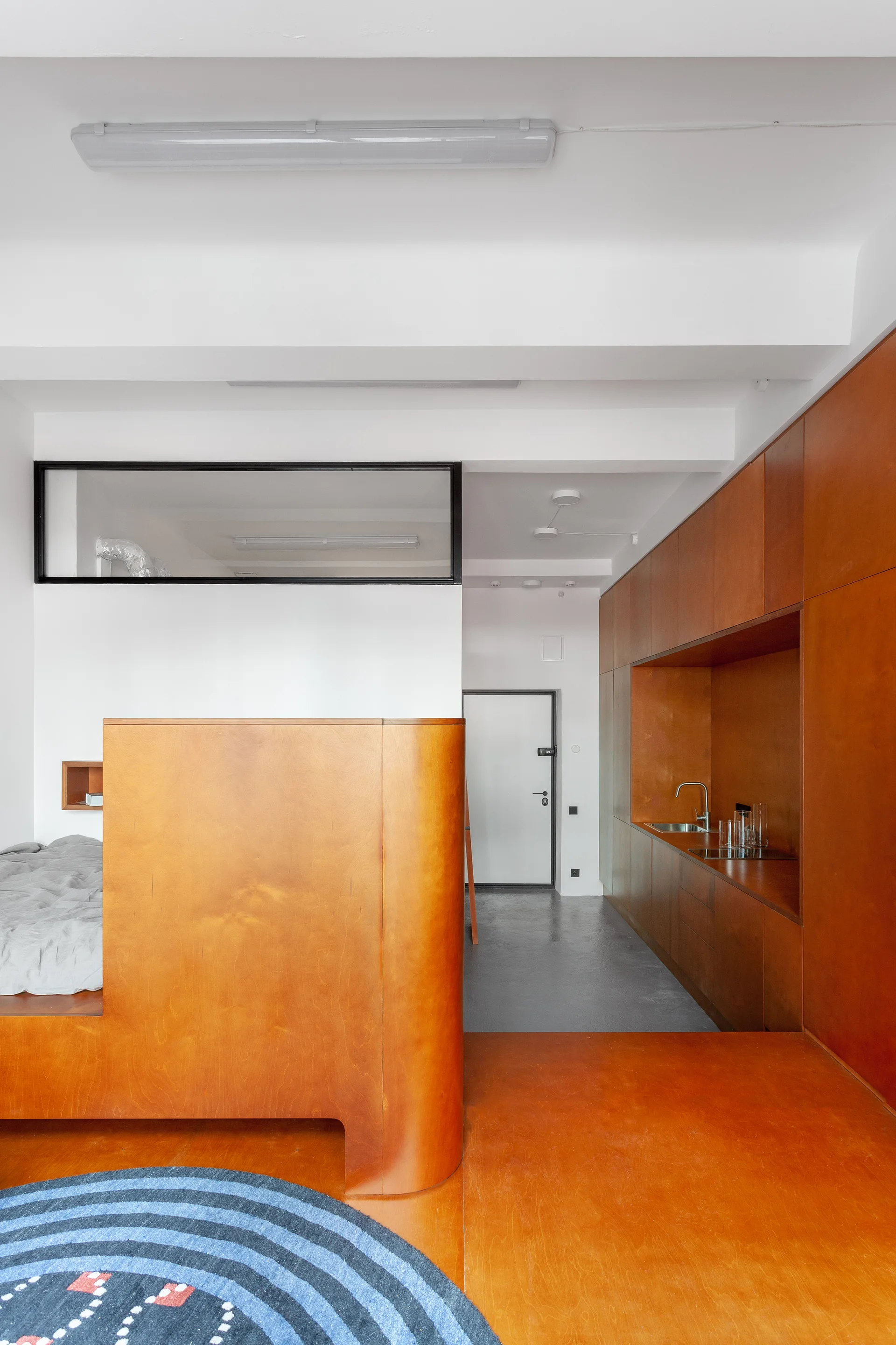 A minimalist studio interior featuring warm orange-toned plywood cabinetry, a curved partition wall, white walls, and a round blue rug.