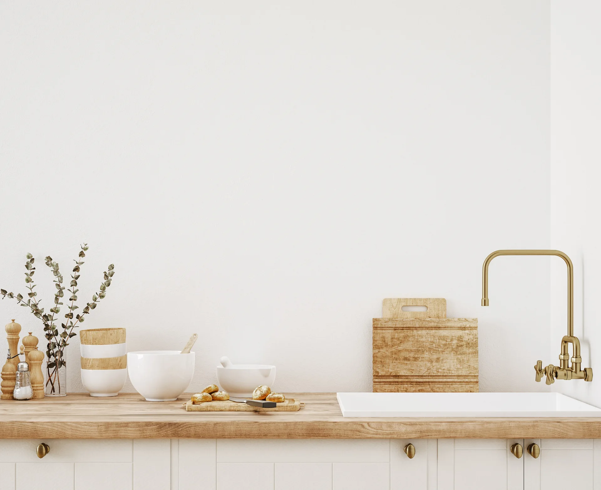 A minimalist white kitchen counter features an undermount sink, a brass faucet, and various wooden kitchen accessories against a plain wall.