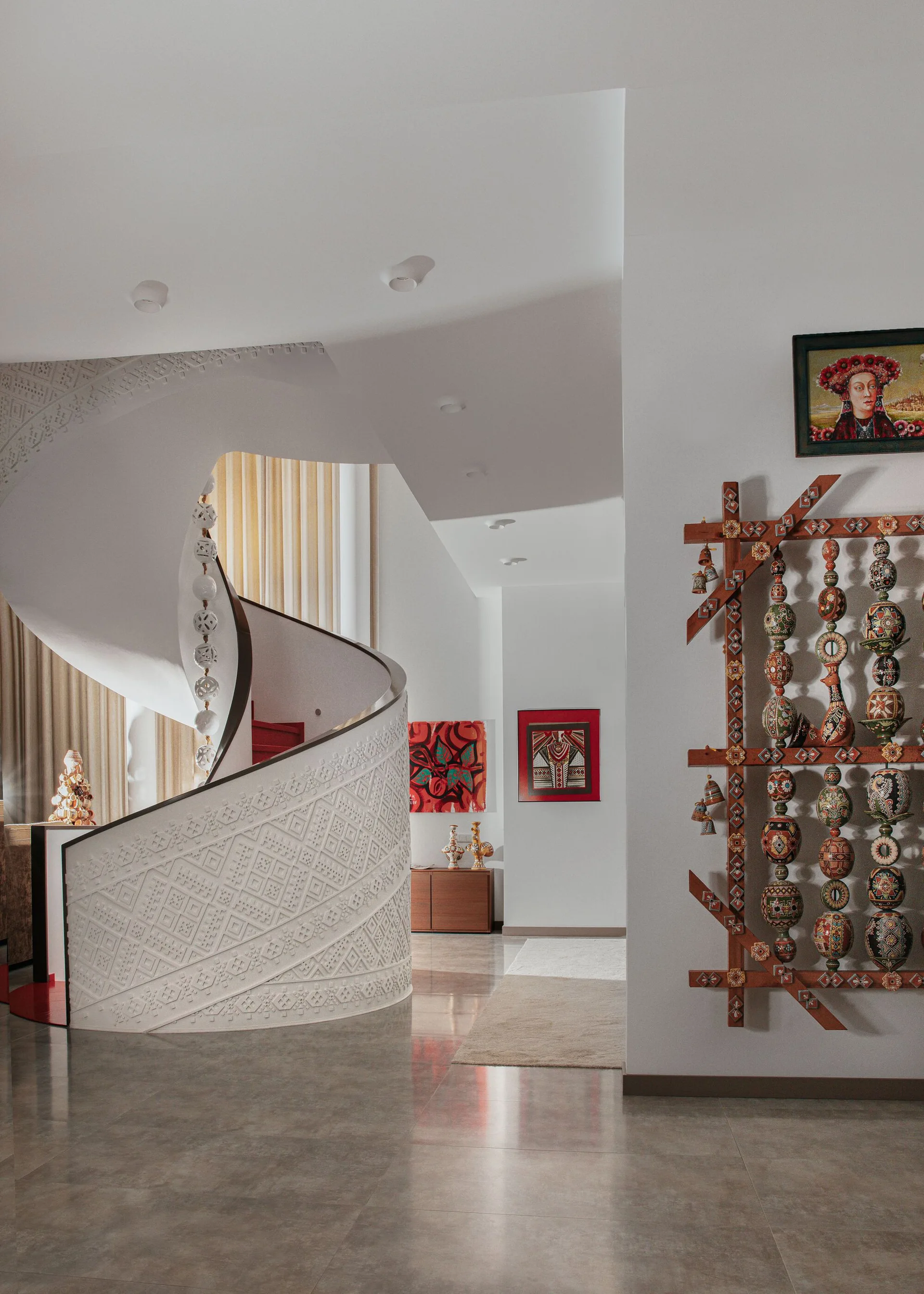 A sculptural white spiral staircase with embossed geometric details stands near a wall decorated with colorful traditional hanging art.