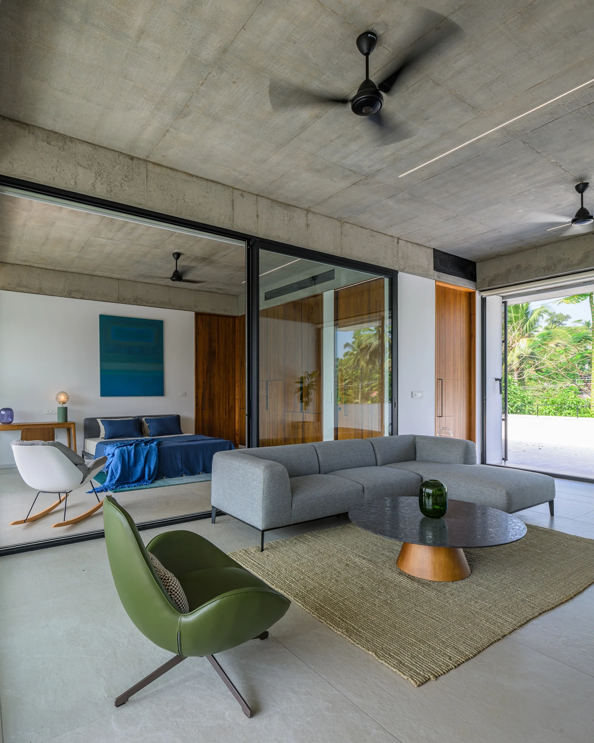 A contemporary living room with a grey sectional, green armchair, and raw concrete ceiling next to a bedroom visible through sliding glass doors.