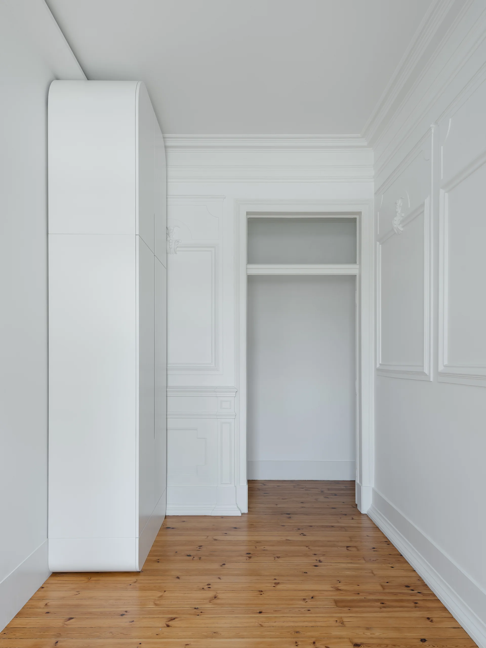 A minimalist white hallway featuring a tall wardrobe with curved edges, classic wall paneling, and warm wood flooring leading to an open doorway.