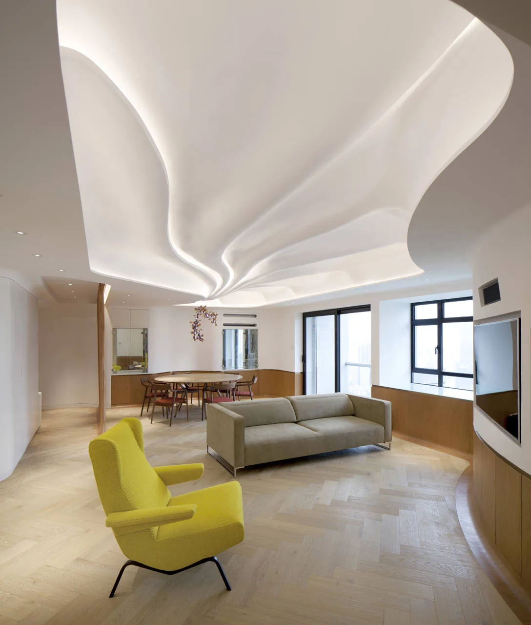 An open-plan living space with a sculptural wave ceiling, a yellow armchair, a beige sofa, and light wood herringbone flooring.