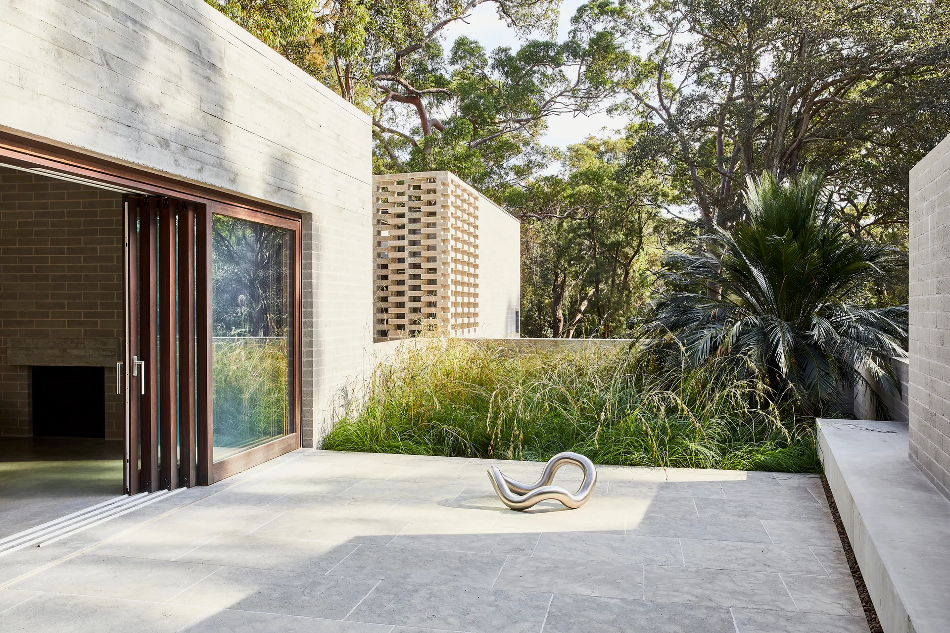A modern concrete patio featuring a bifold wood-framed glass door, a metal sculpture on the stone tile floor, and wild grass landscaping.