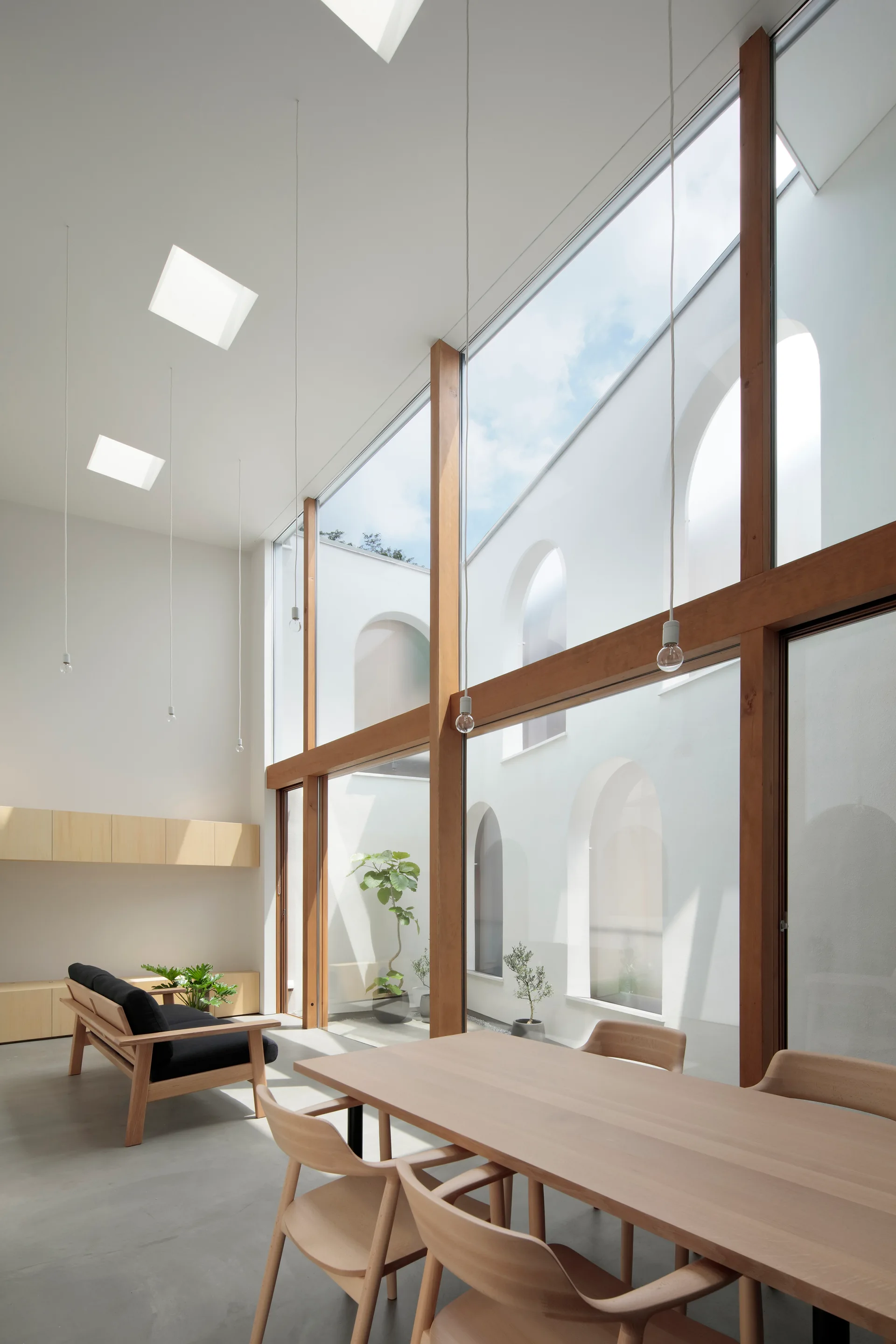 A minimalist open-plan dining and living room features a light wood table, a black sofa, and double-height wood-framed windows facing a white courtyard.