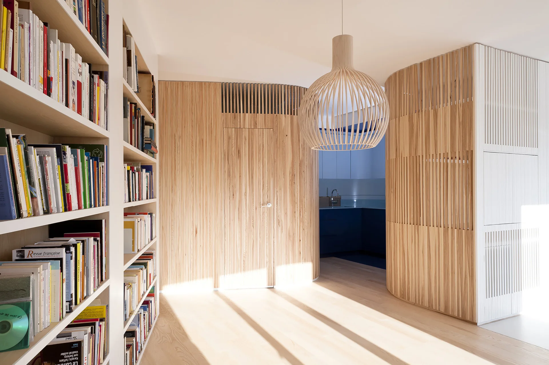A bright hallway features curved vertical wood-slat walls, a matching spherical pendant light, and tall white built-in bookshelves loaded with books.