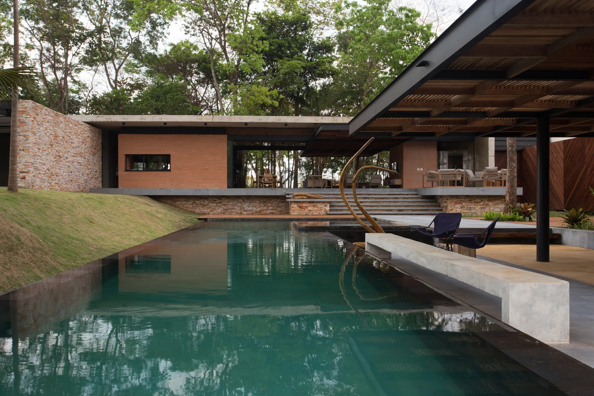 A contemporary outdoor swimming pool features a long concrete bench, stone retaining walls, and a pavilion with a wood slatted ceiling.