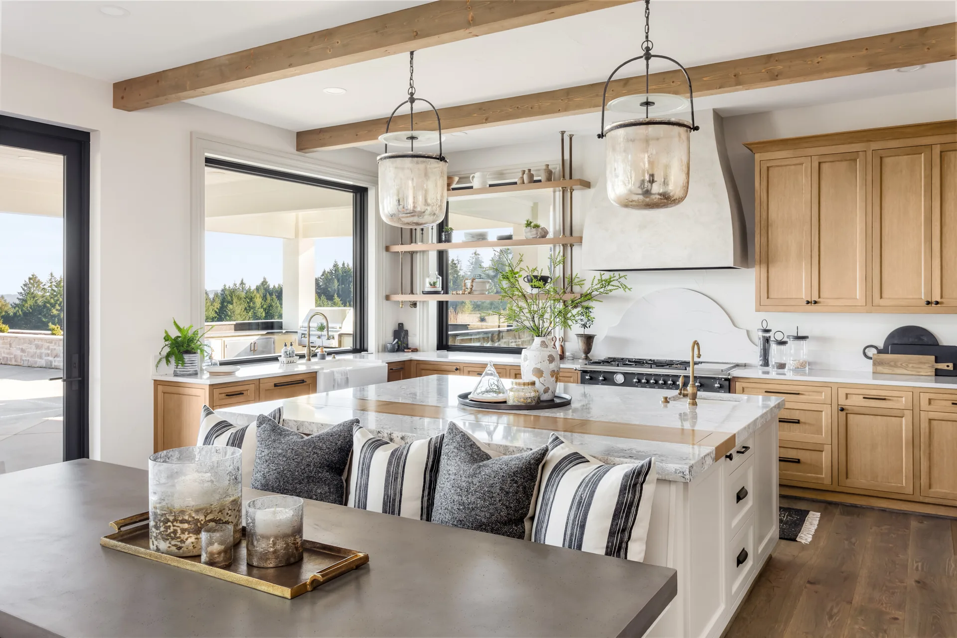 A spacious modern farmhouse kitchen featuring a central island, light wood cabinetry, exposed ceiling beams, and large glass pendant lights.