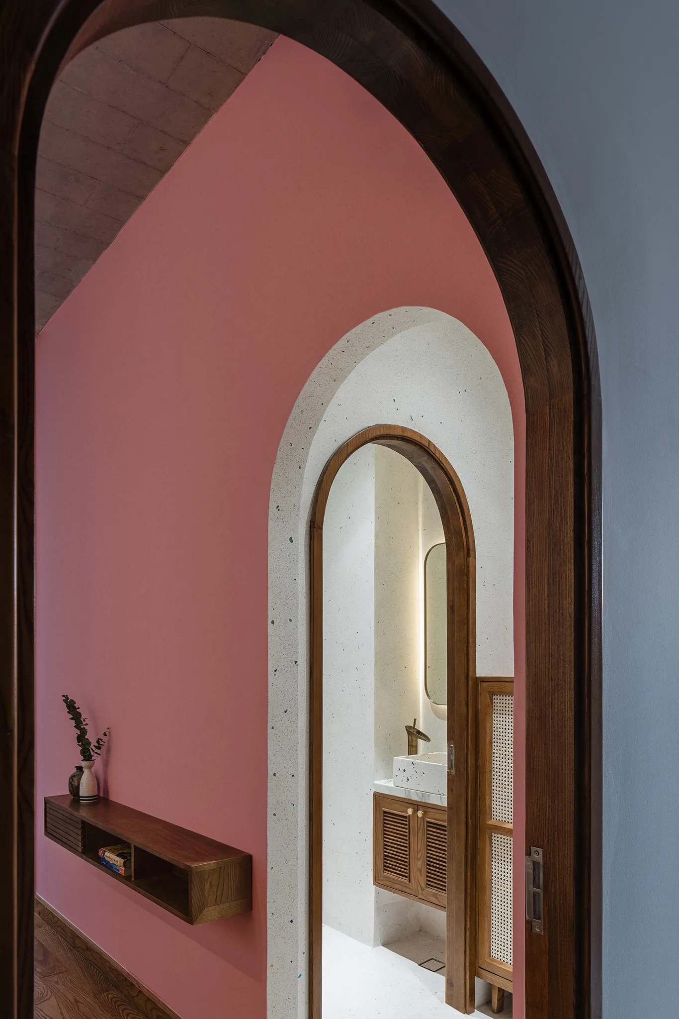 A view through nested arches with pink walls and a wooden console shelf leading toward a bathroom vanity with a backlit oval mirror.