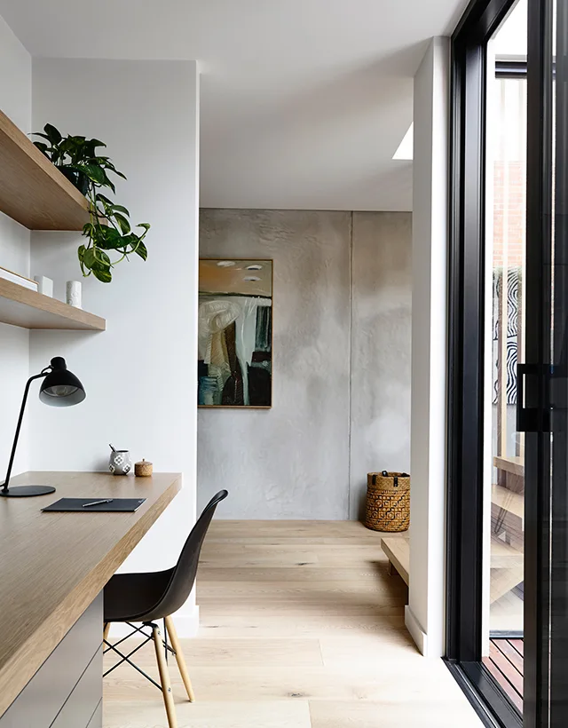 A minimalist home office features a wooden built-in desk with a black desk lamp, floating shelves, and a textured grey plaster wall in the background.