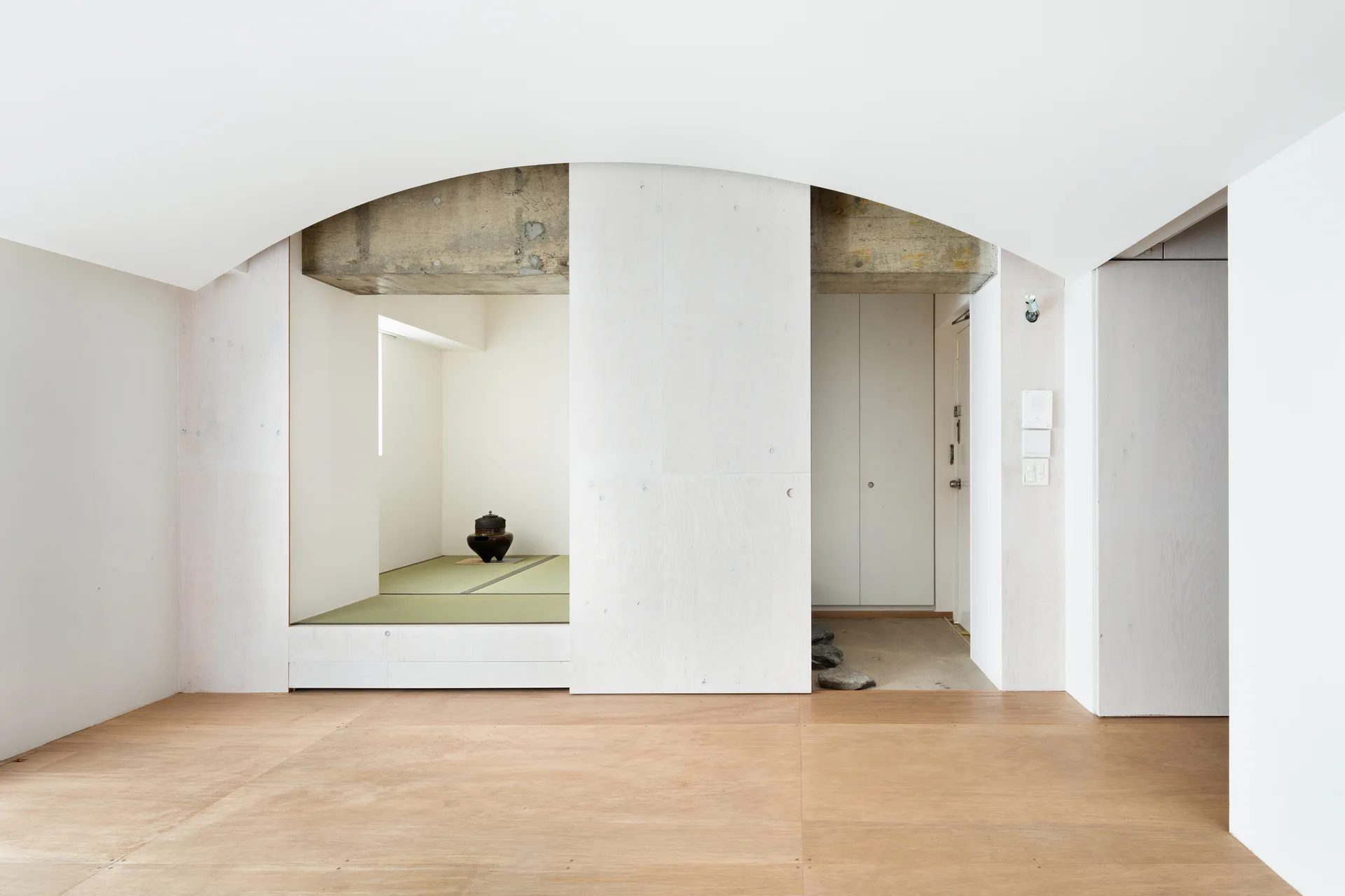 A minimalist interior featuring a raised tatami room with green mats behind a white arched ceiling and exposed concrete beams.
