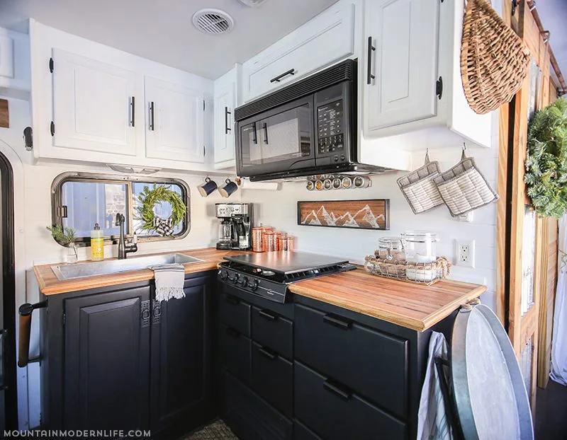 A small kitchen with matte black base cabinets, white upper cabinets, warm wood countertops, and a black microwave.