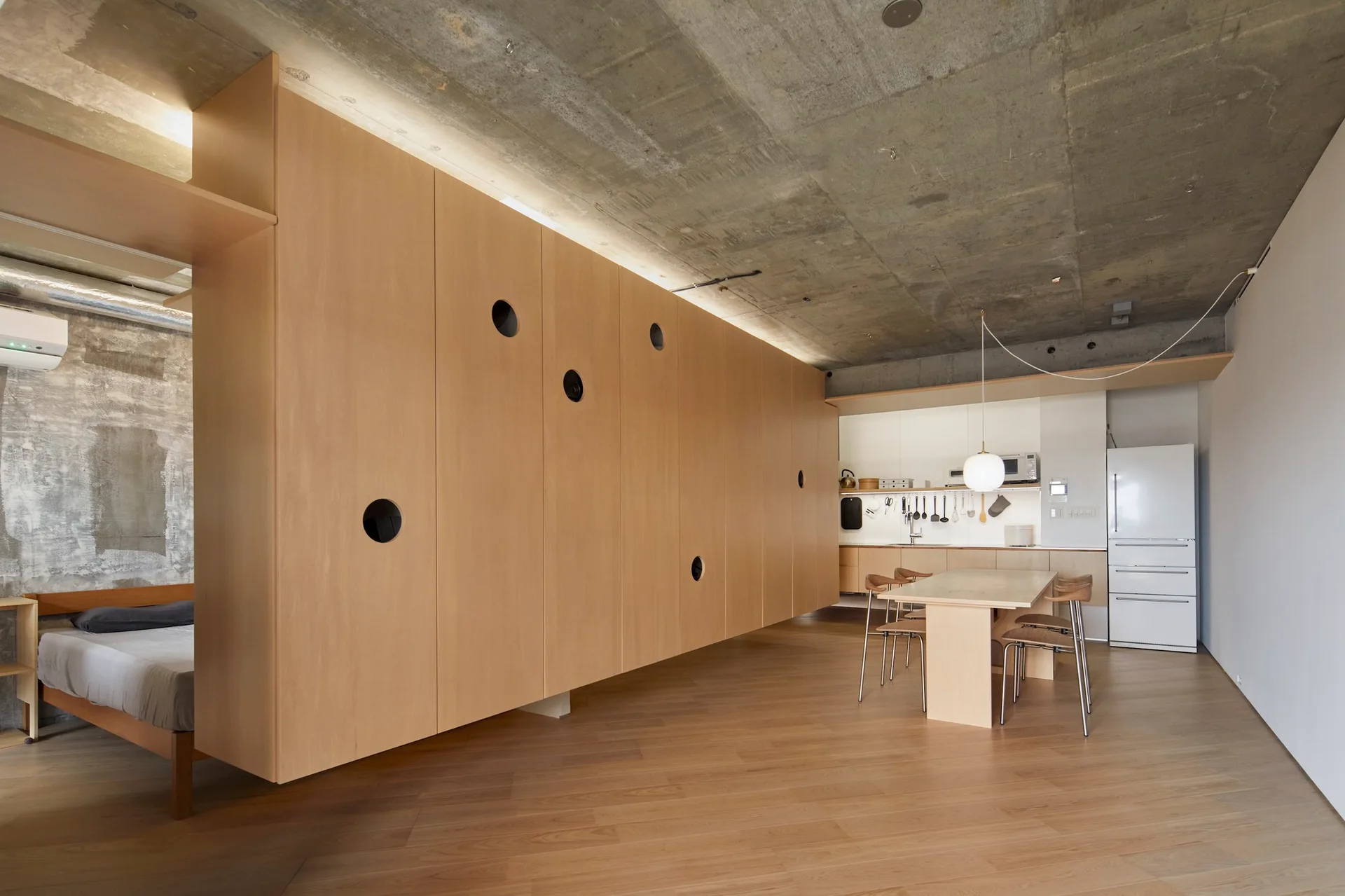 A large wooden storage partition with circular cutout handles separates a bedroom from a dining space under an exposed concrete ceiling.