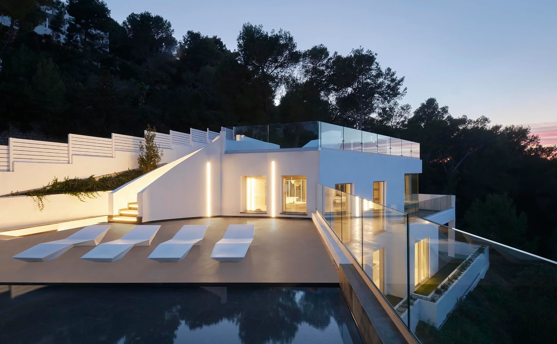 A modern white stucco house terrace features sculptural lounge chairs beside a swimming pool, enclosed by glass railings at twilight.