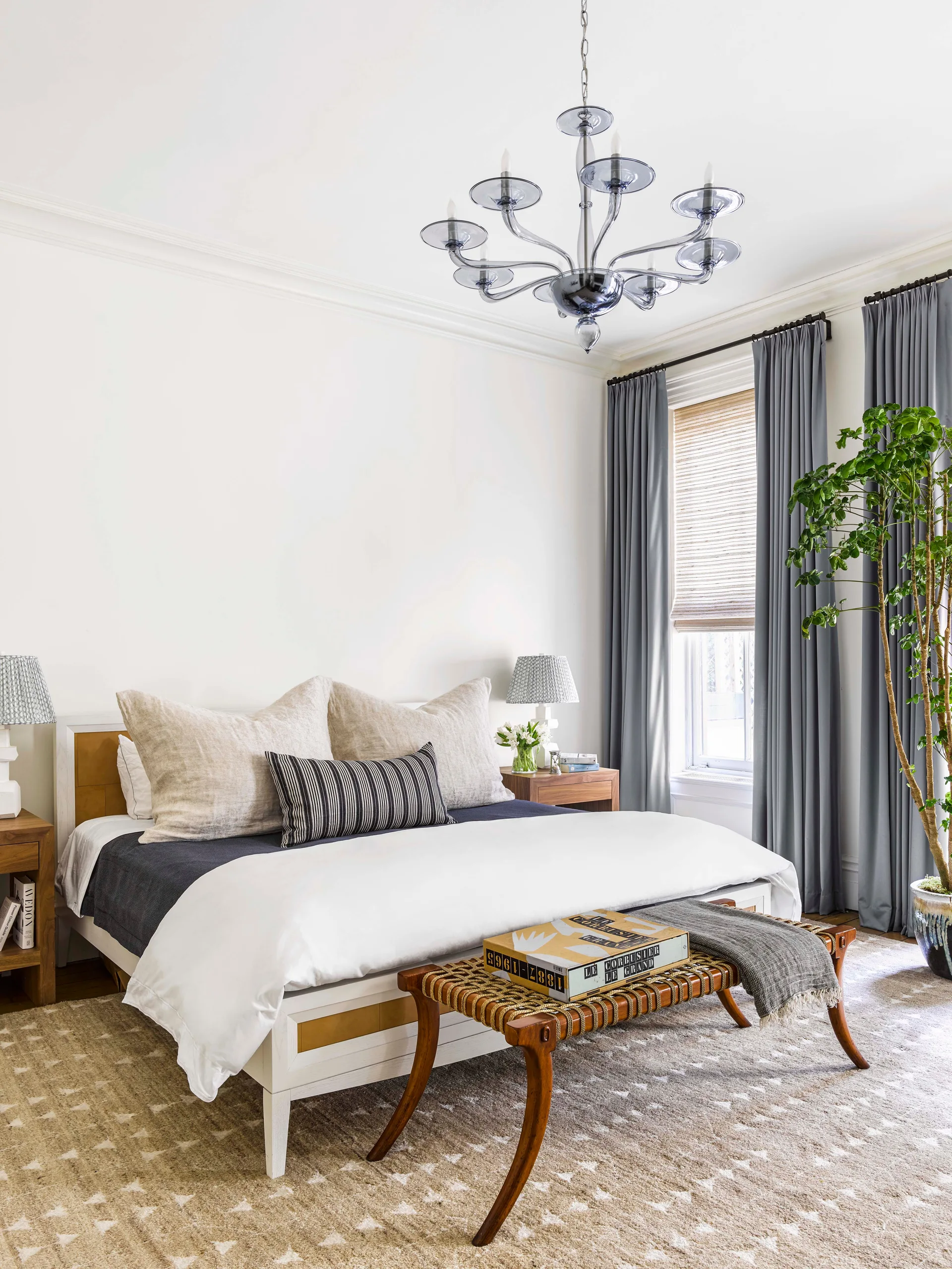 A bright transitional bedroom features a central bed with white linens, flanked by wooden nightstands, a woven bench, and long grey curtains.