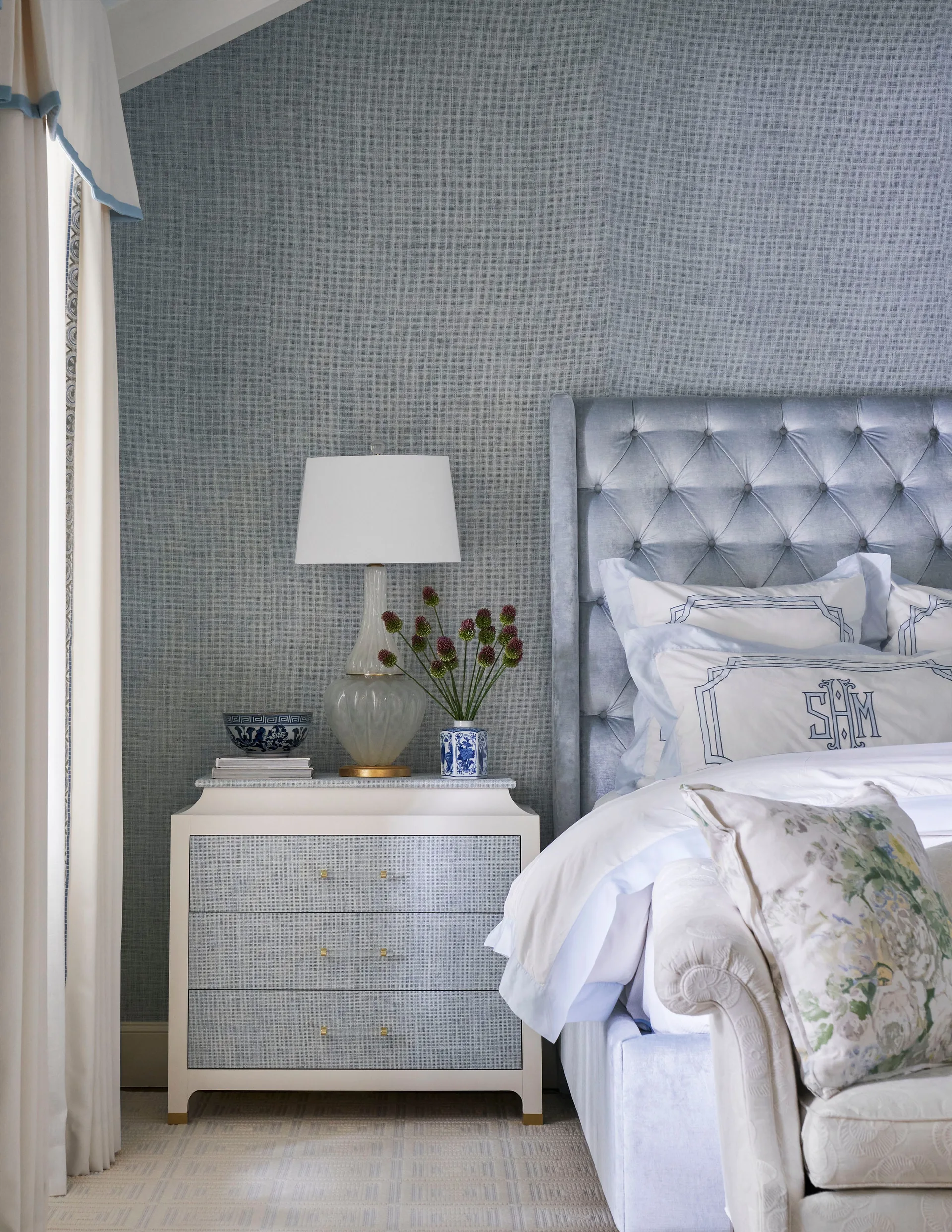 A bedroom featuring a blue tufted headboard, a white nightstand with fabric-front drawers, a glass lamp, and textured blue wallpaper.