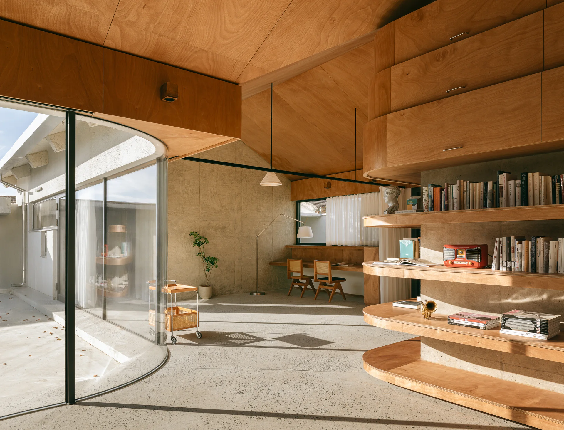 A modern home office with a sloped plywood ceiling, curved wooden shelving, exposed concrete walls, and a large curved glass wall.