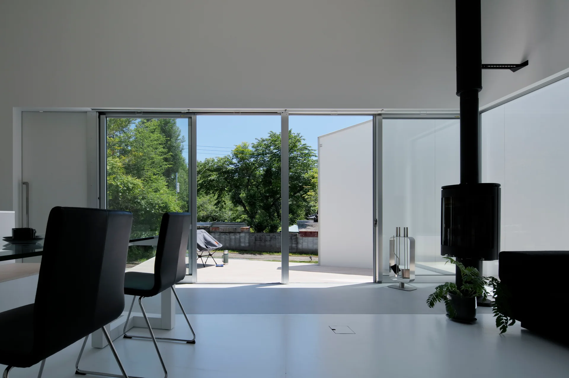 A minimalist white living room features black dining chairs, a black freestanding fireplace, and large sliding glass doors opening to a sunny outdoor patio.