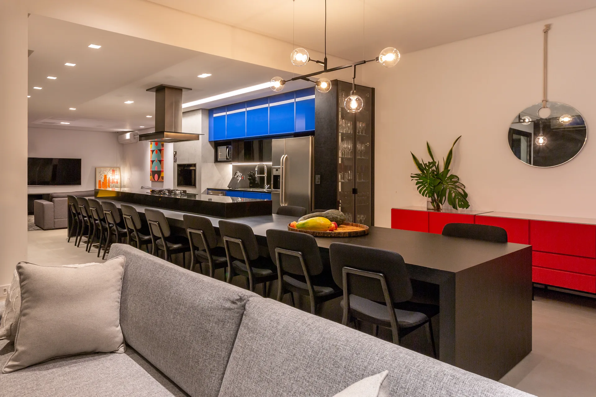 An open-concept kitchen features bright blue cabinets, a long black dining table with chairs, a grey sofa, and a vibrant red sideboard.