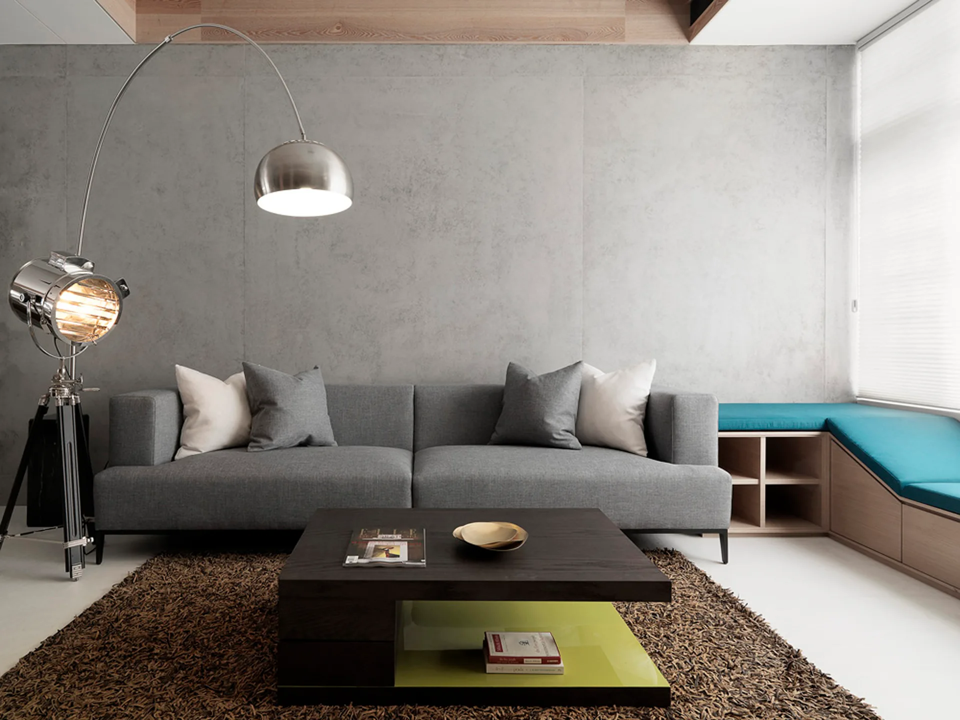 A grey modern sofa stands against a grey concrete wall next to a dark wood coffee table, a brown shag rug, and a large metal arc lamp.