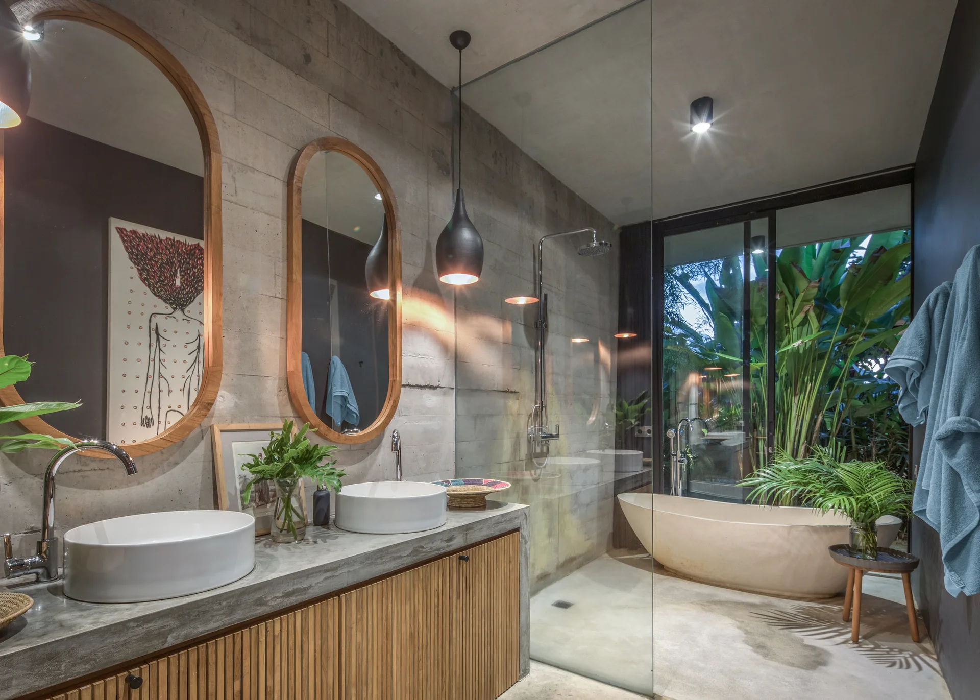 A concrete bathroom features a double vessel sink vanity with wood slatted cabinets, oval mirrors, and a glass-enclosed shower with a freestanding tub.