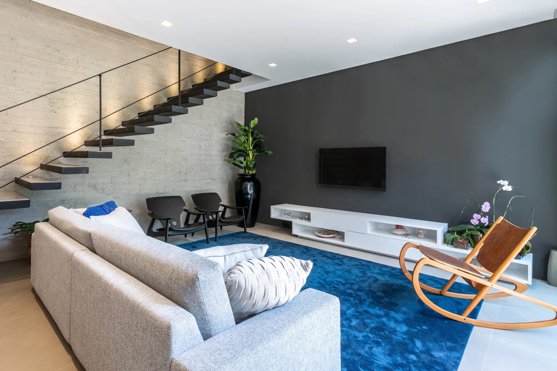 A contemporary living room features a grey sofa on a blue rug, a low white media console against a dark wall, and a black floating staircase.