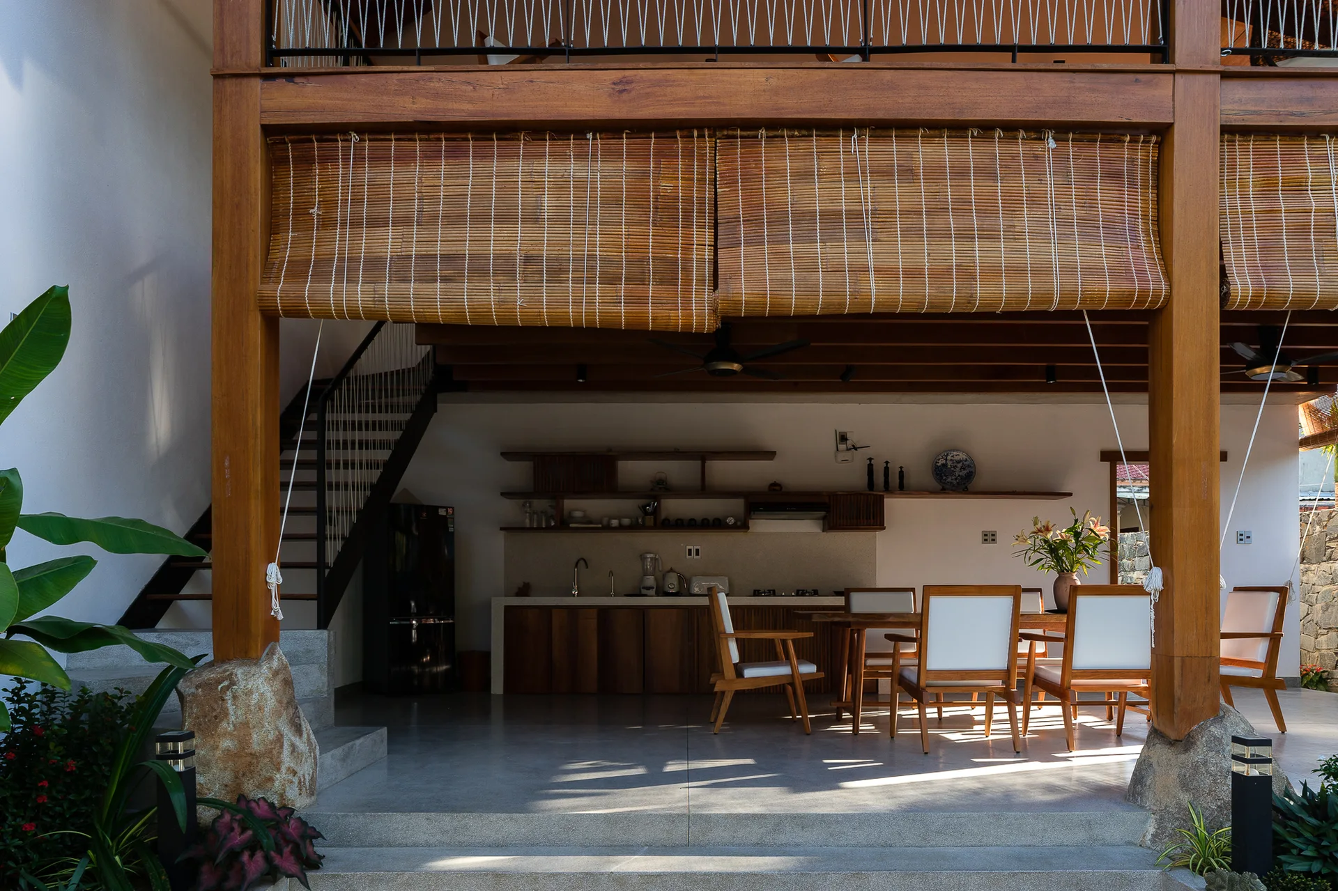 An open-air tropical patio featuring a wooden dining set, a simple outdoor kitchen, concrete flooring, and rolled bamboo blinds hanging from timber beams.