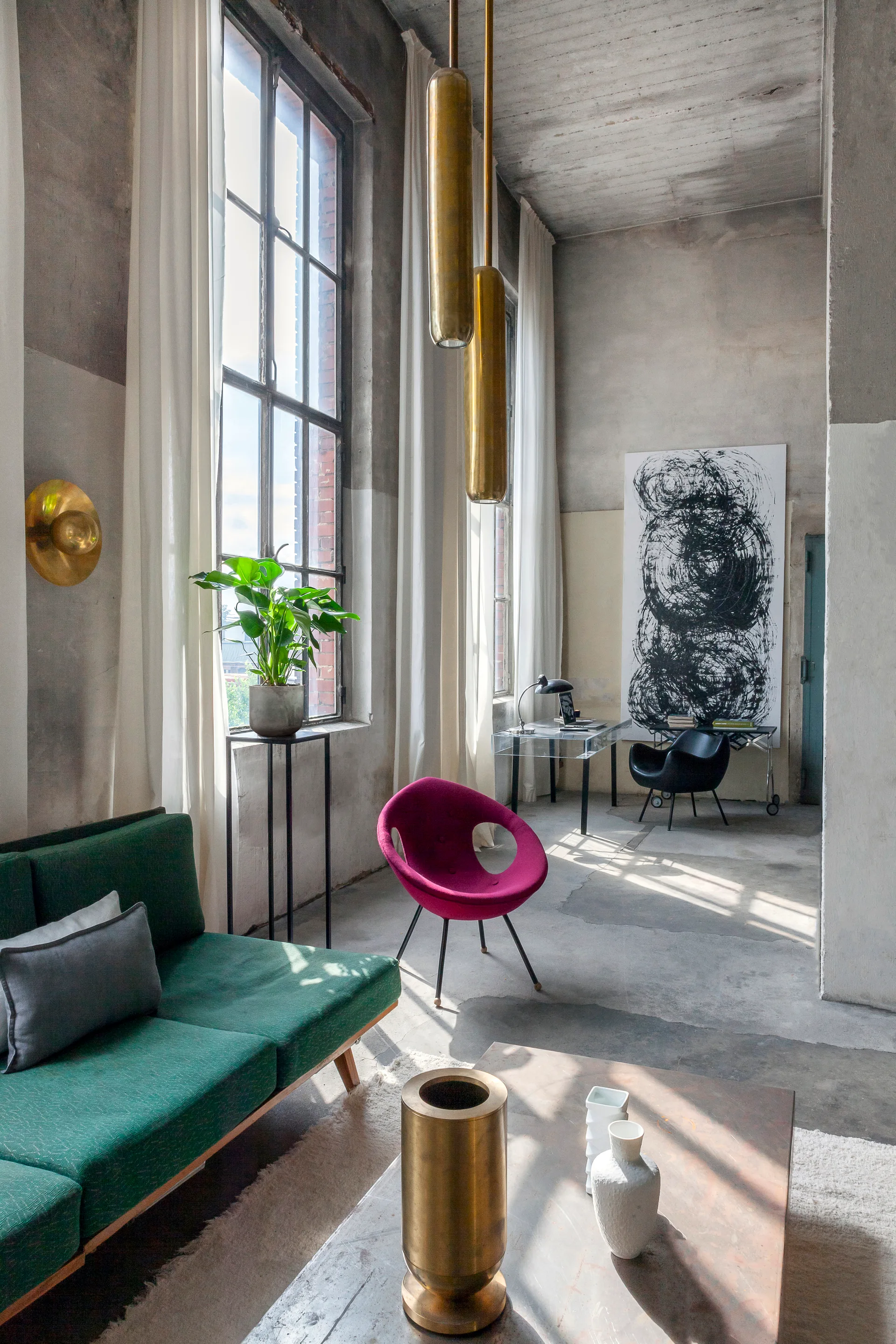 An industrial loft living room features tall windows, a green sofa, a magenta chair, and cylindrical brass pendant lights.