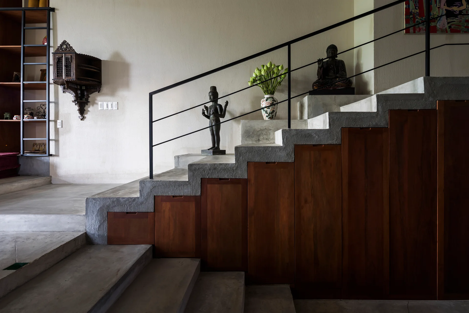 A concrete staircase with a black metal railing features integrated wooden storage cabinets underneath and decorative statues resting on the steps.