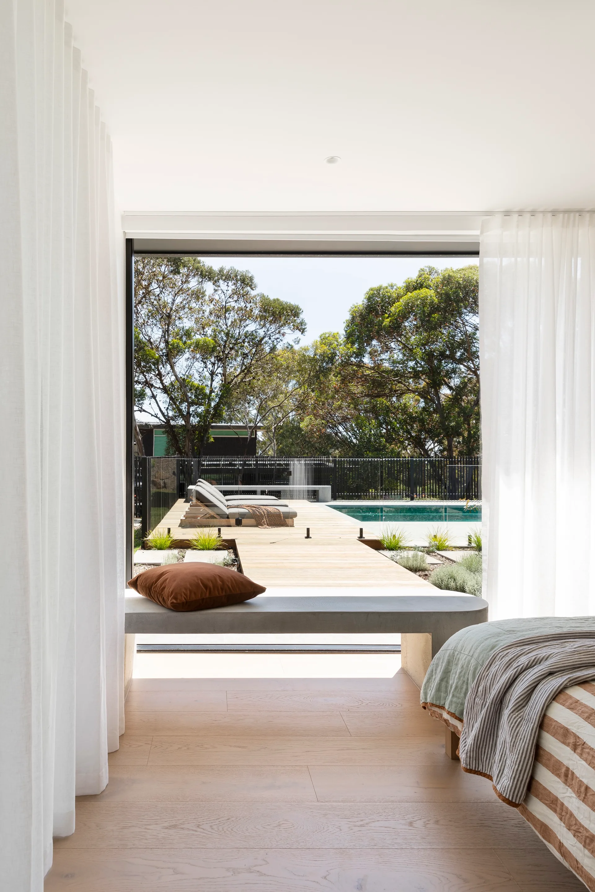 A light-filled bedroom features a concrete window bench, sheer white curtains, and a sliding glass door opening to an outdoor wood deck with a pool.