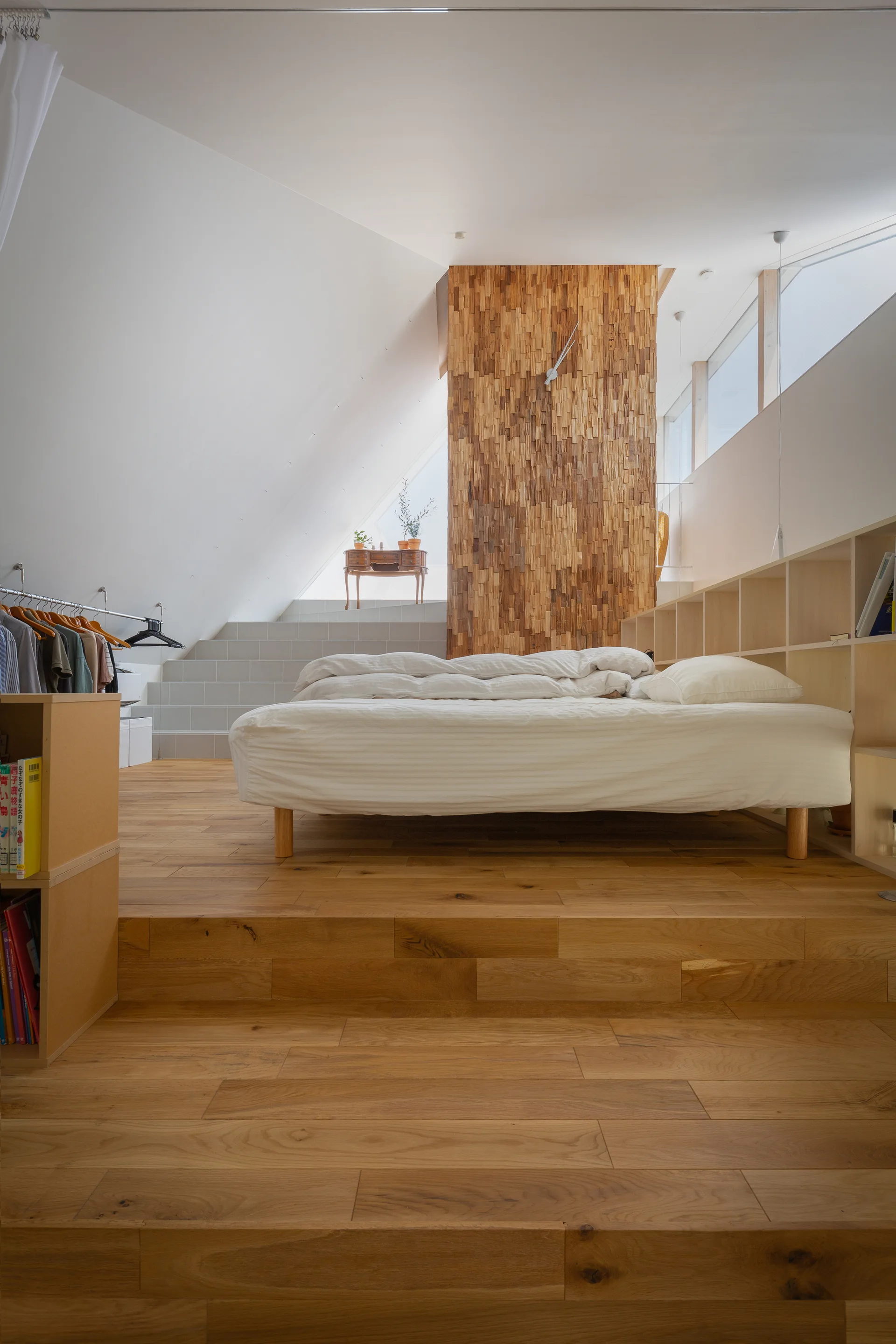 A loft bedroom features a platform bed on tiered wood flooring next to a textured wood-plank accent wall and built-in light wood shelving.