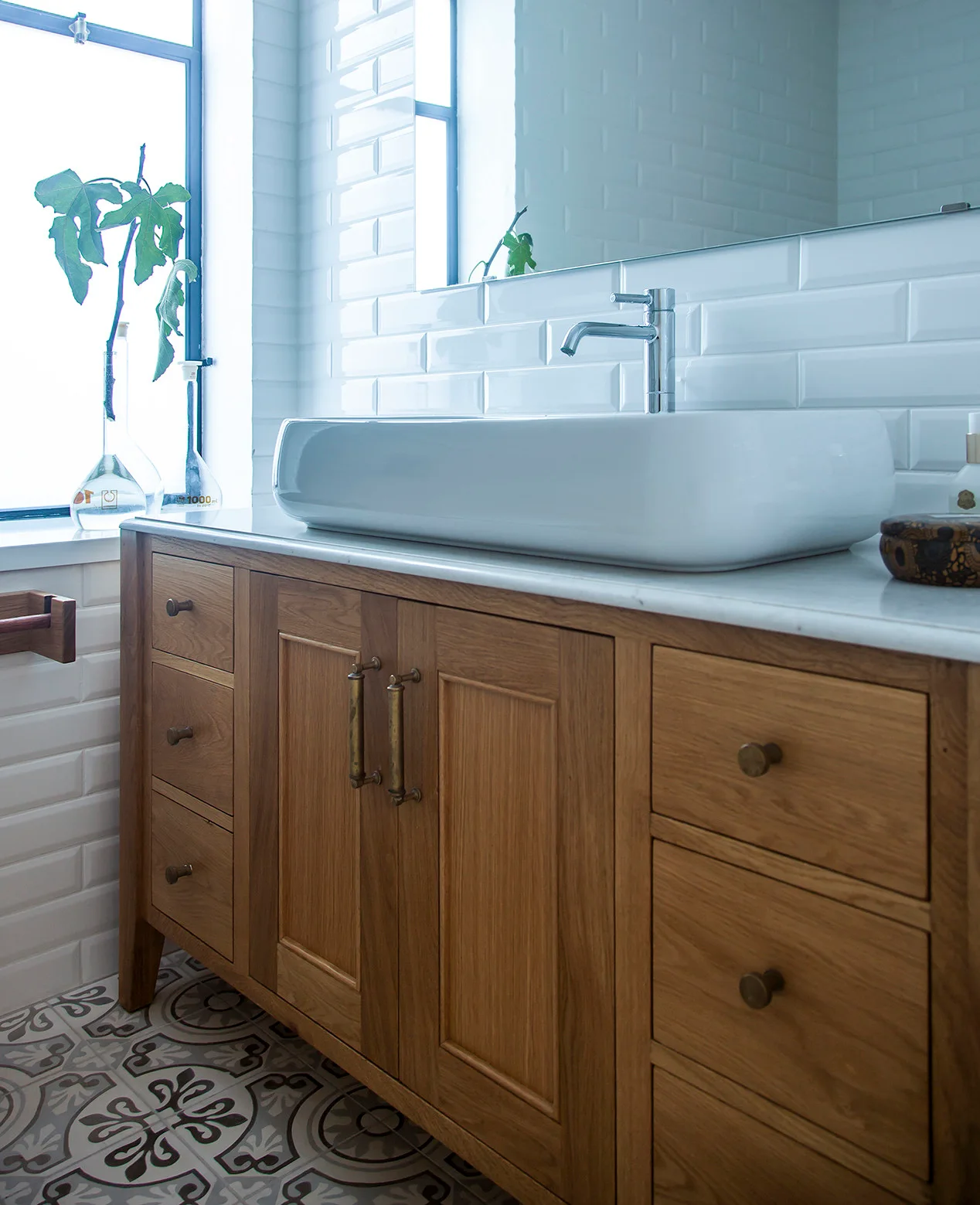 A wooden bathroom vanity with a marble countertop and white vessel sink stands against a white subway tile wall and patterned floor.