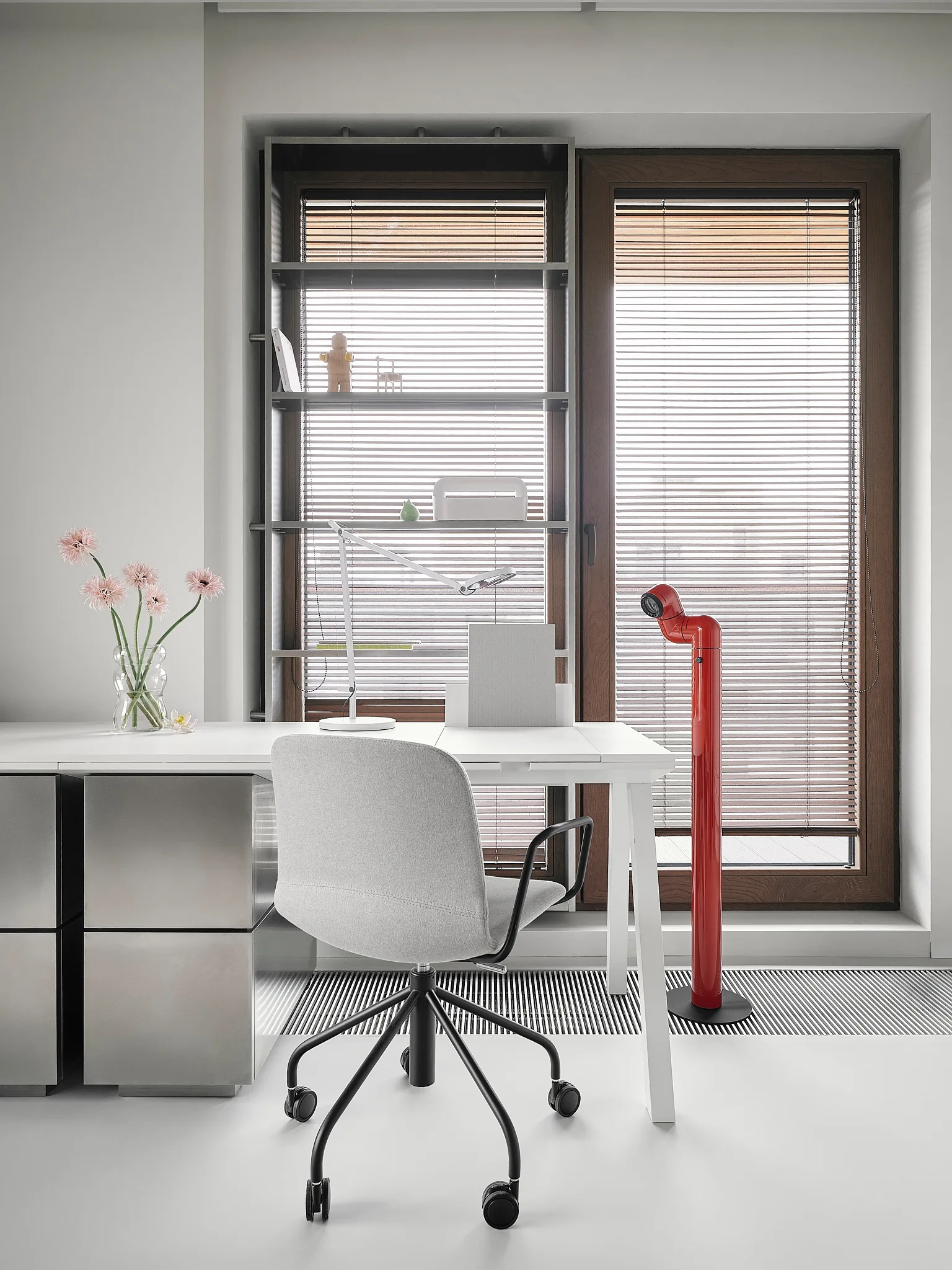 Minimalist workspace with a white desk, grey fabric chair, brushed metal drawers, and a red industrial floor lamp positioned by a window.