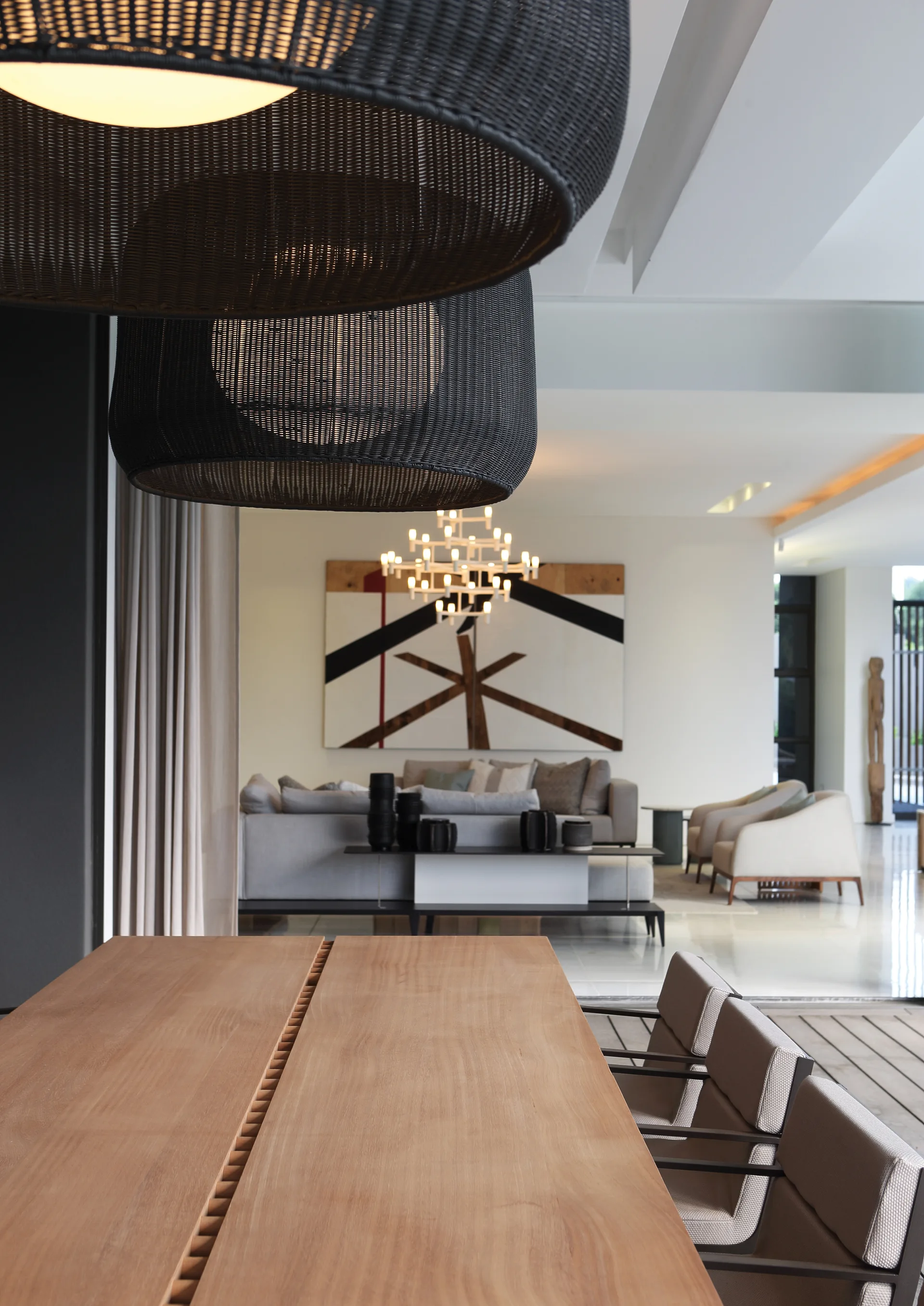 A modern open-concept dining and living space featuring a long wooden table, dark woven pendant lights, a gray sofa, and geometric wall art.