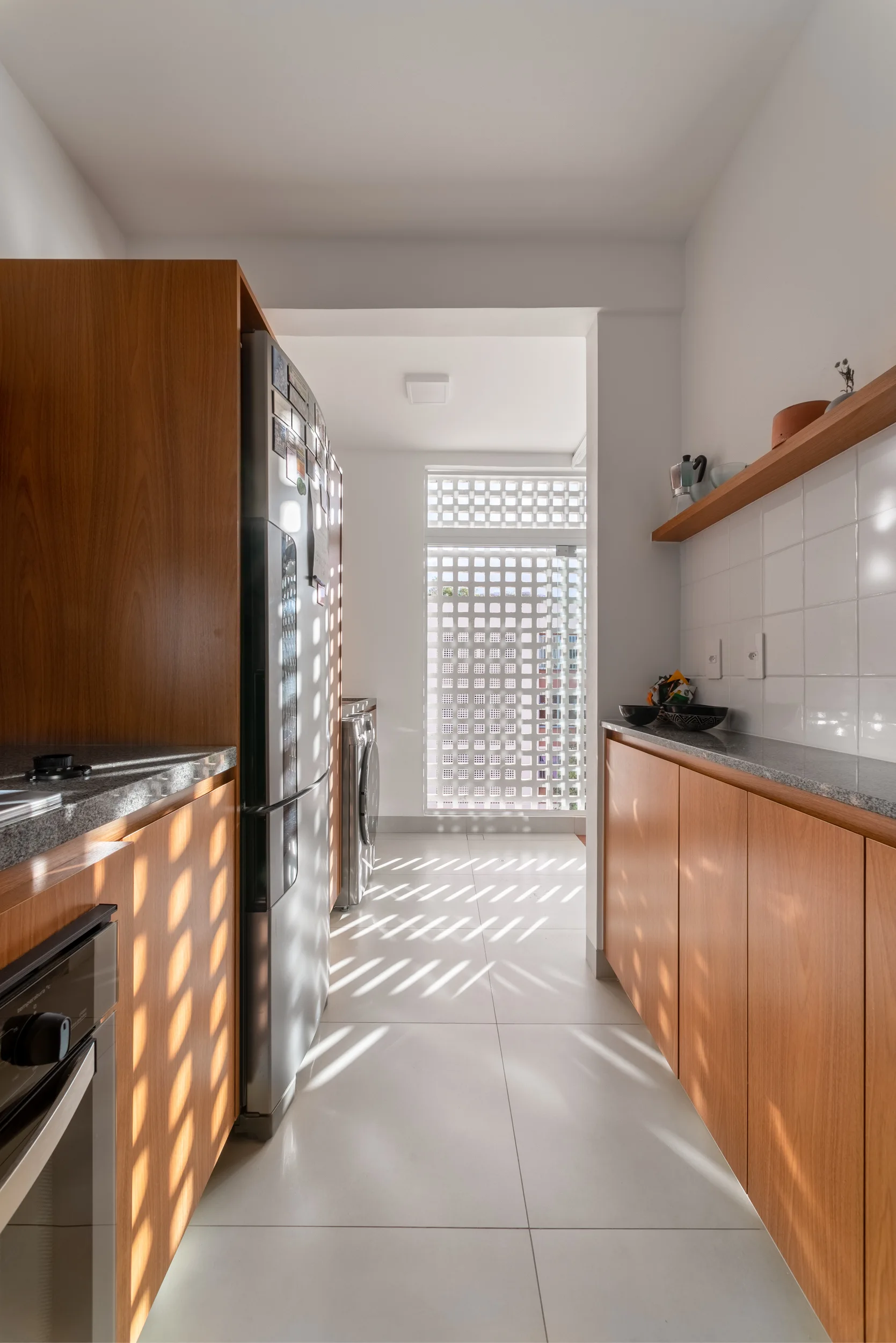 A narrow galley kitchen features wood cabinets, a stainless steel refrigerator, and a geometric screen window casting grid-patterned light on the tile floor.