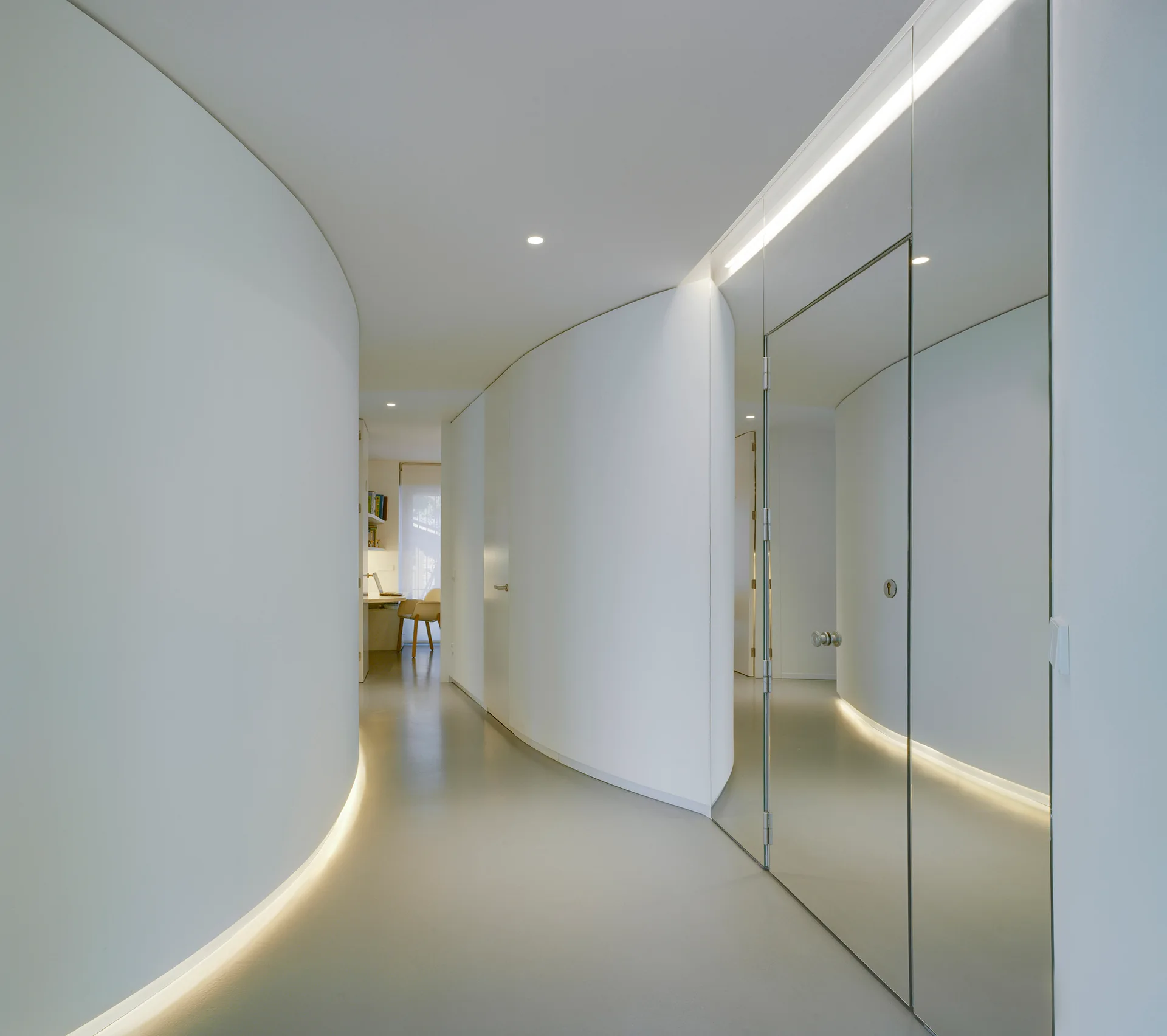 A minimalist white hallway featuring a sweeping curved wall, floor-level strip lighting, and tall mirrored doors reflecting the corridor.