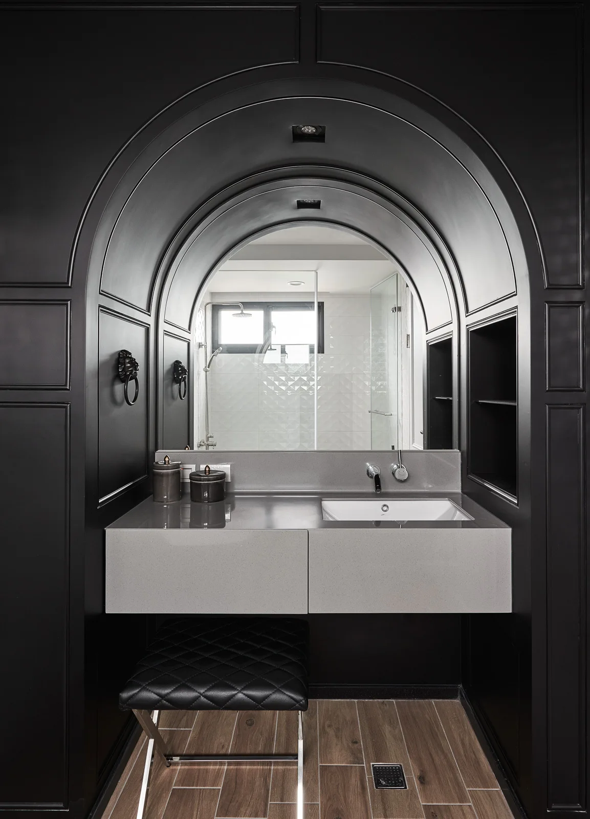 A bathroom vanity with a grey floating sink set into a matte black arched paneled wall, featuring an integrated mirror and a quilted stool below.