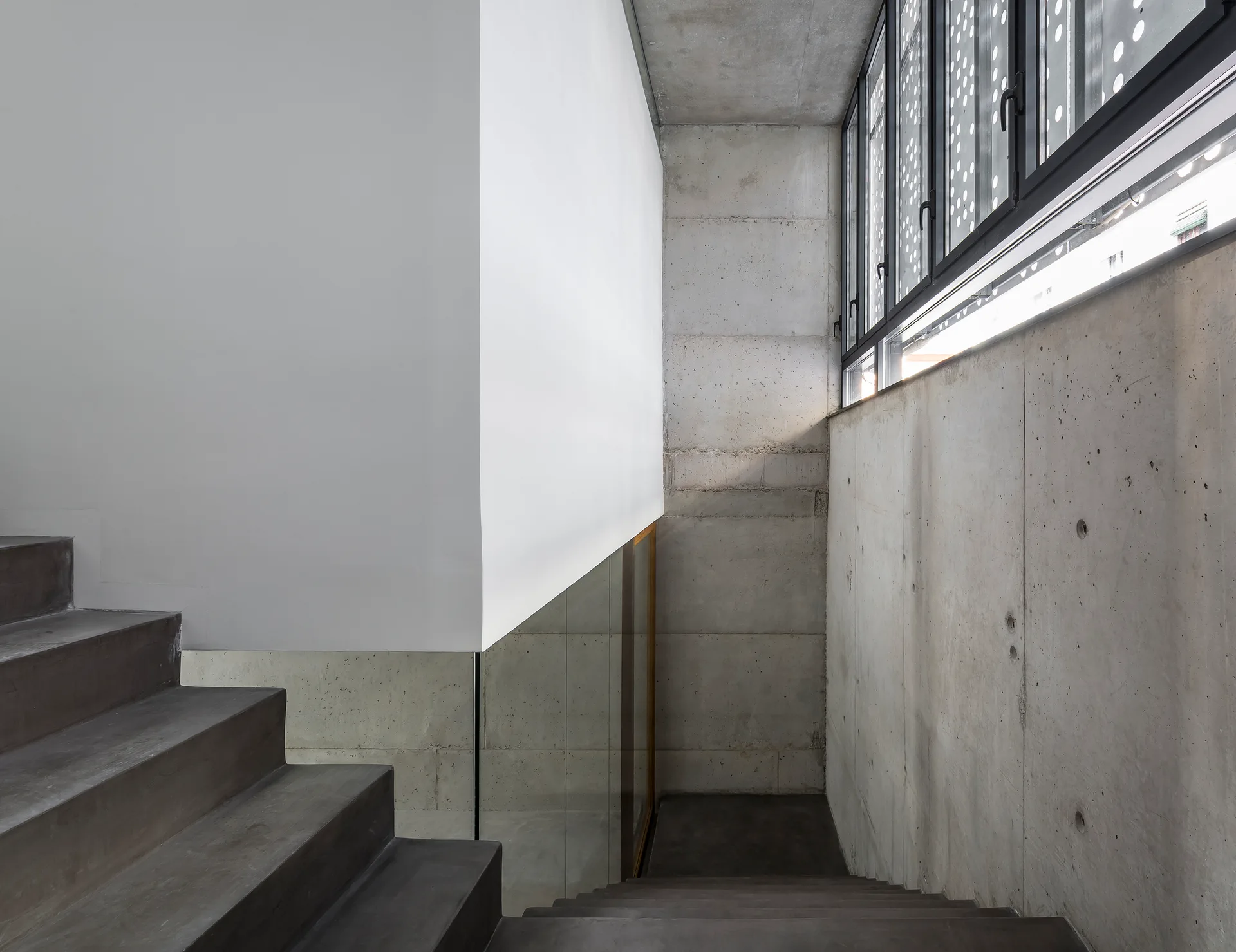 A minimalist stairwell featuring dark concrete steps, raw concrete walls, a large suspended white volume, and industrial metal-framed windows.