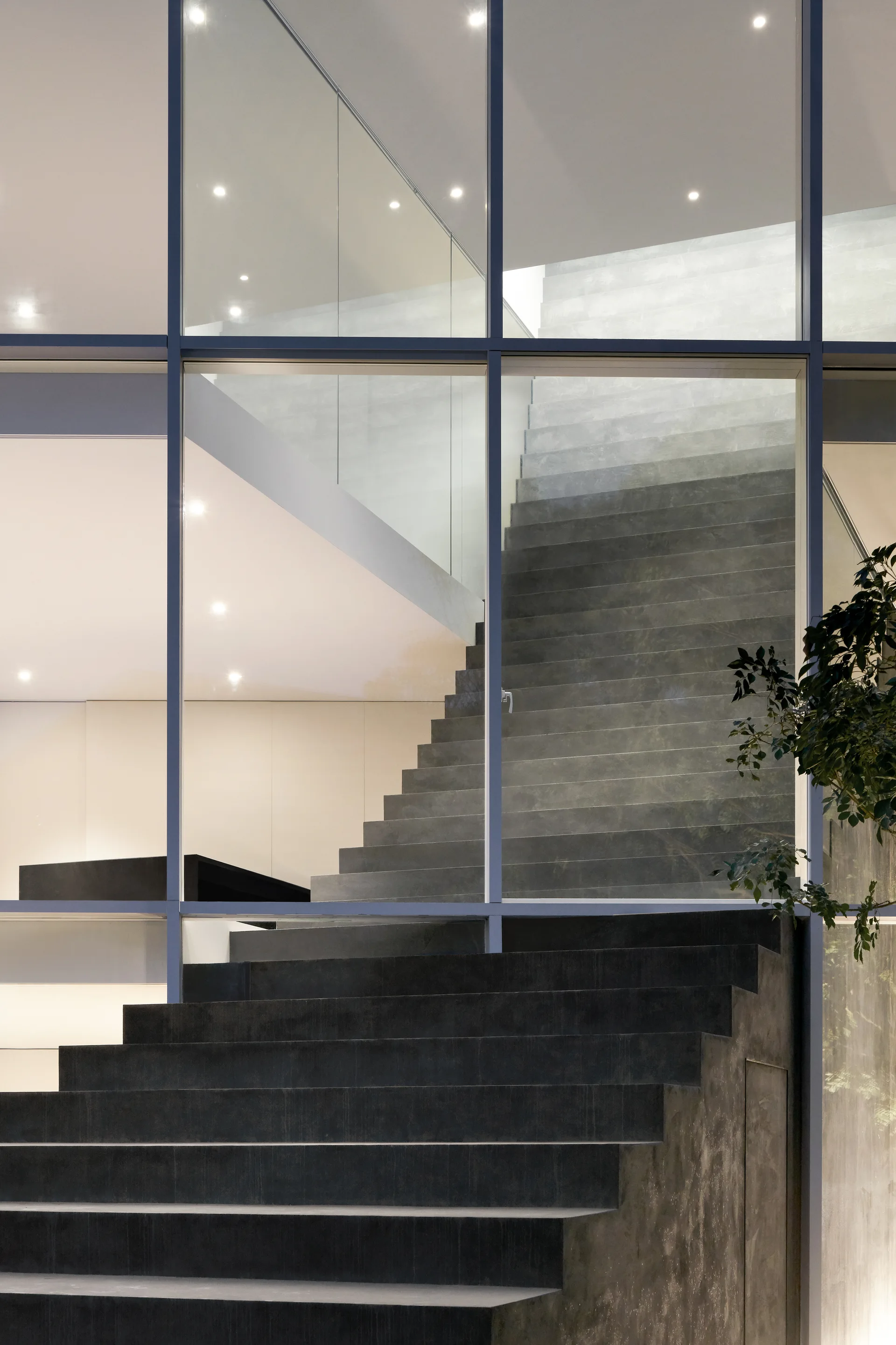 A dark grey stone staircase rises behind a large floor-to-ceiling glass grid facade lit by recessed ceiling spotlights.