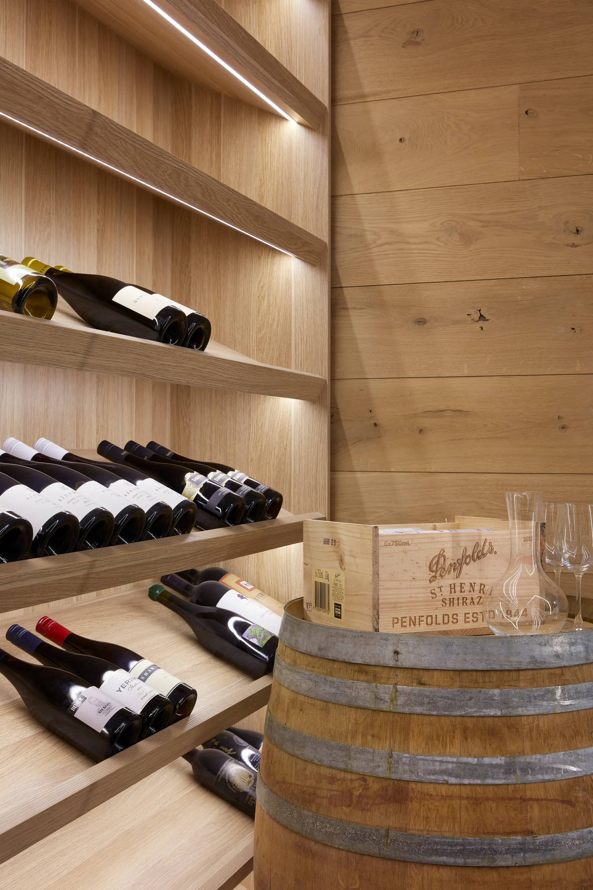 Custom light wood shelving displays wine bottles under integrated LED lighting next to an oak barrel topped with a wooden crate and decanter.