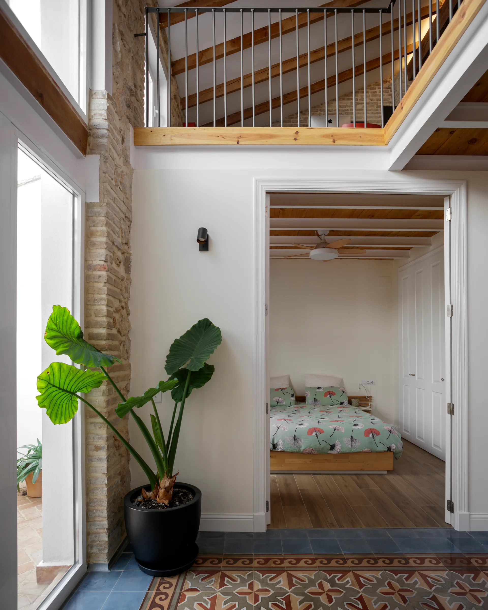 A view through a white doorway into a bedroom, featuring patterned floor tiles, a large potted plant, exposed brick, and a mezzanine balcony overhead.