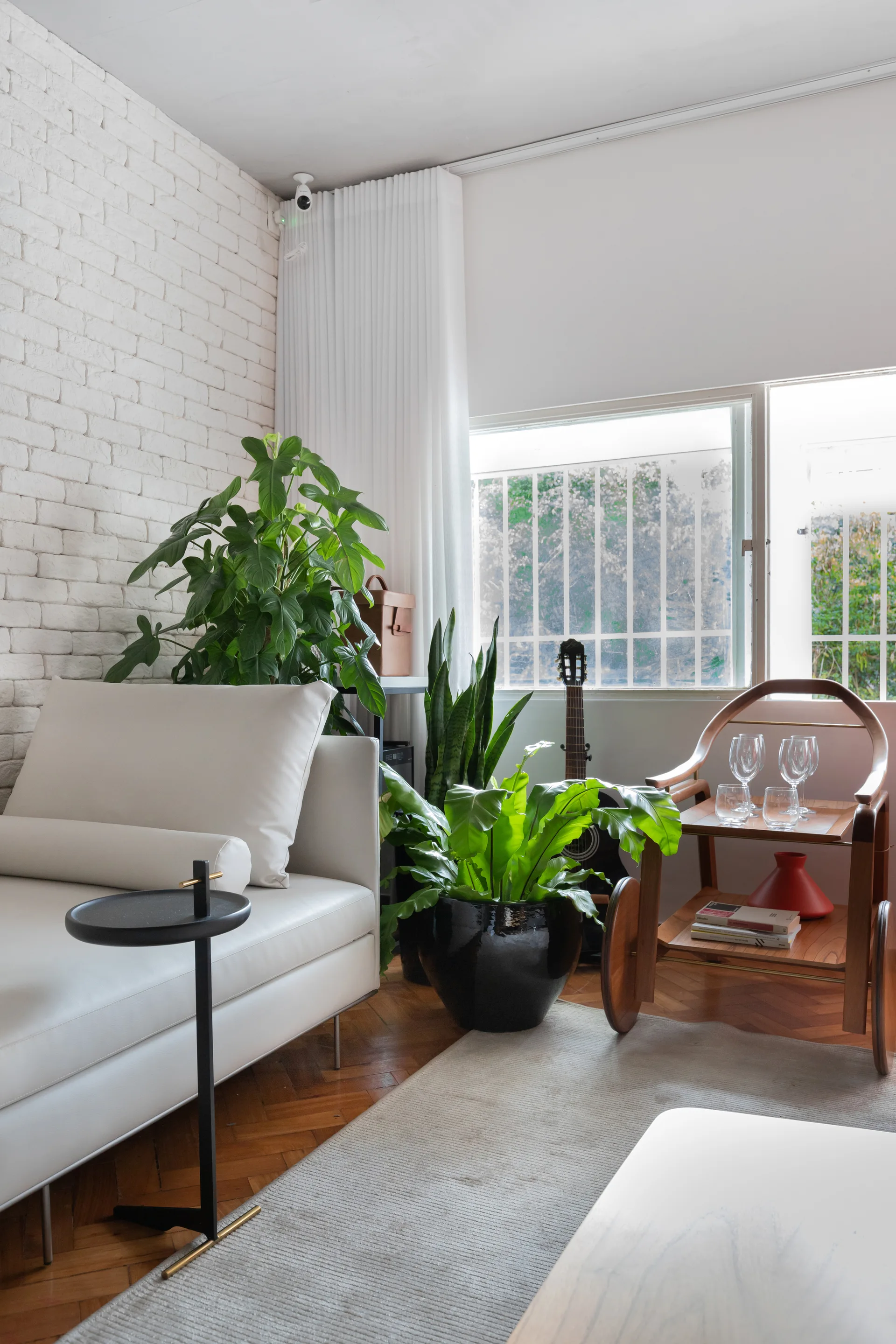 A white sofa and a wooden bar cart sit on herringbone flooring in a room with a painted white brick wall and potted green plants.