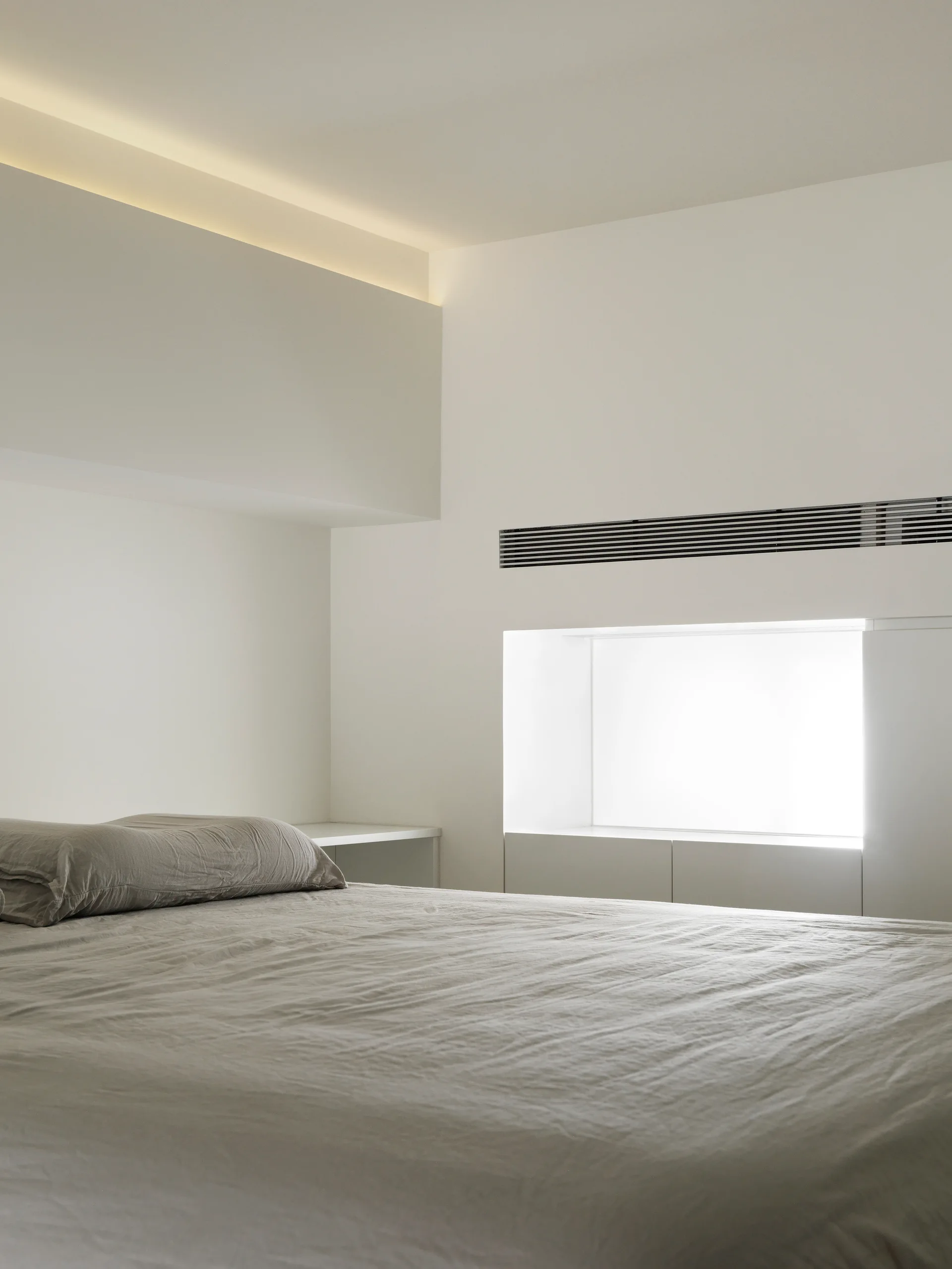 A minimalist white bedroom features a bed with grey linens beneath a ceiling with warm cove lighting and a deep recessed window.