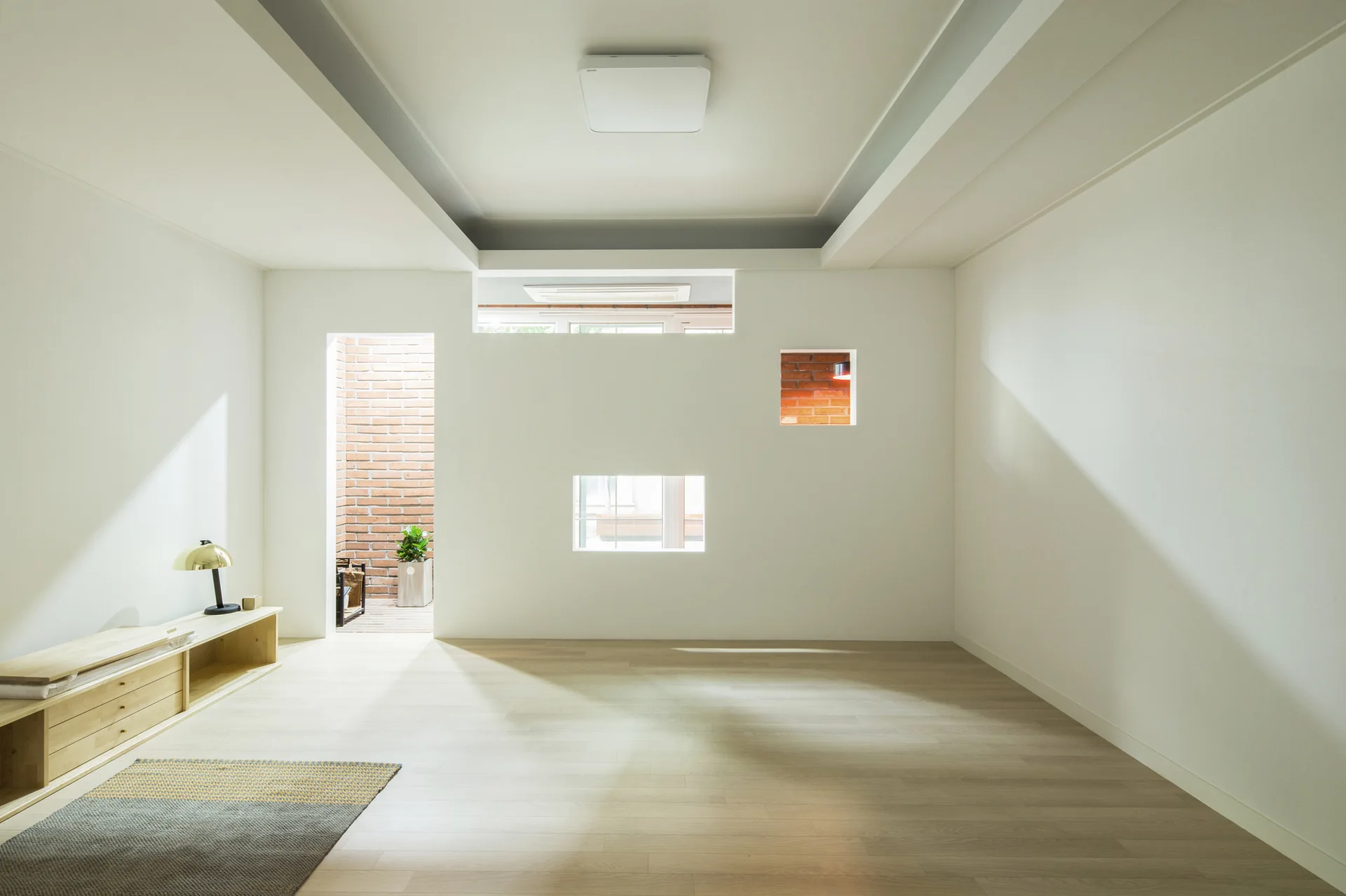 A minimalist white living room features light wood floors, a low media console, and a central wall with several square cutout window openings.
